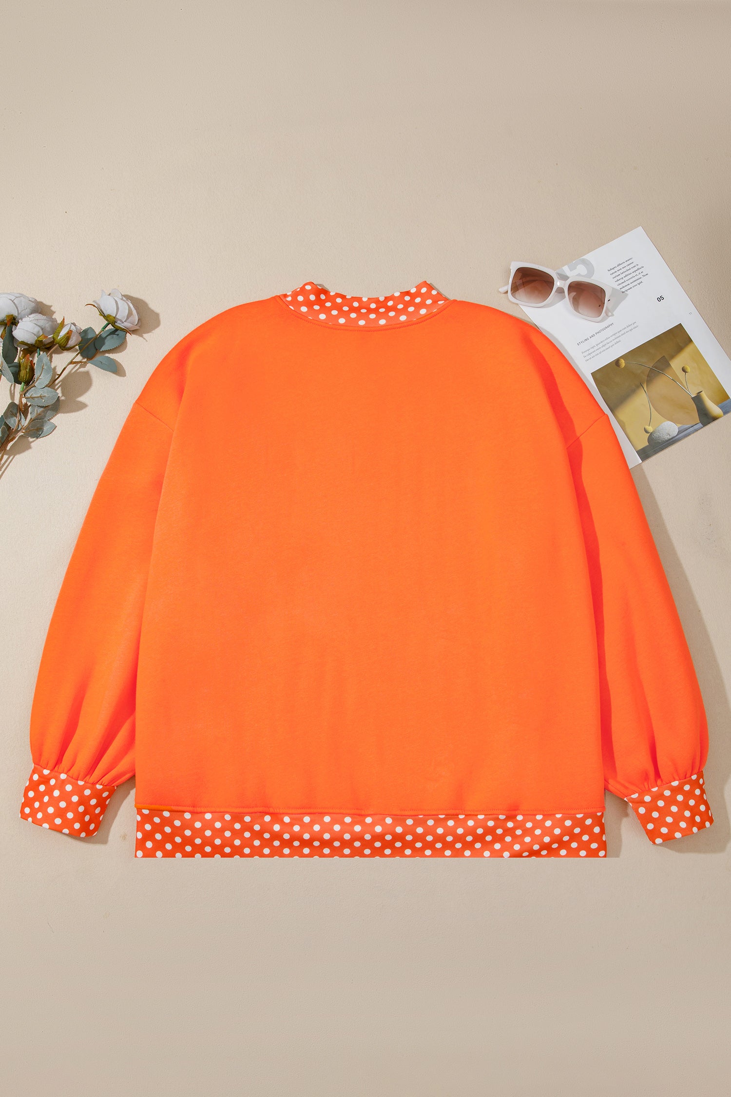 Bright orange long-sleeve top with white polka dot trim at the crew neck, cuffs, and hem, laid flat on a beige background with decorative sunglasses and flowers.