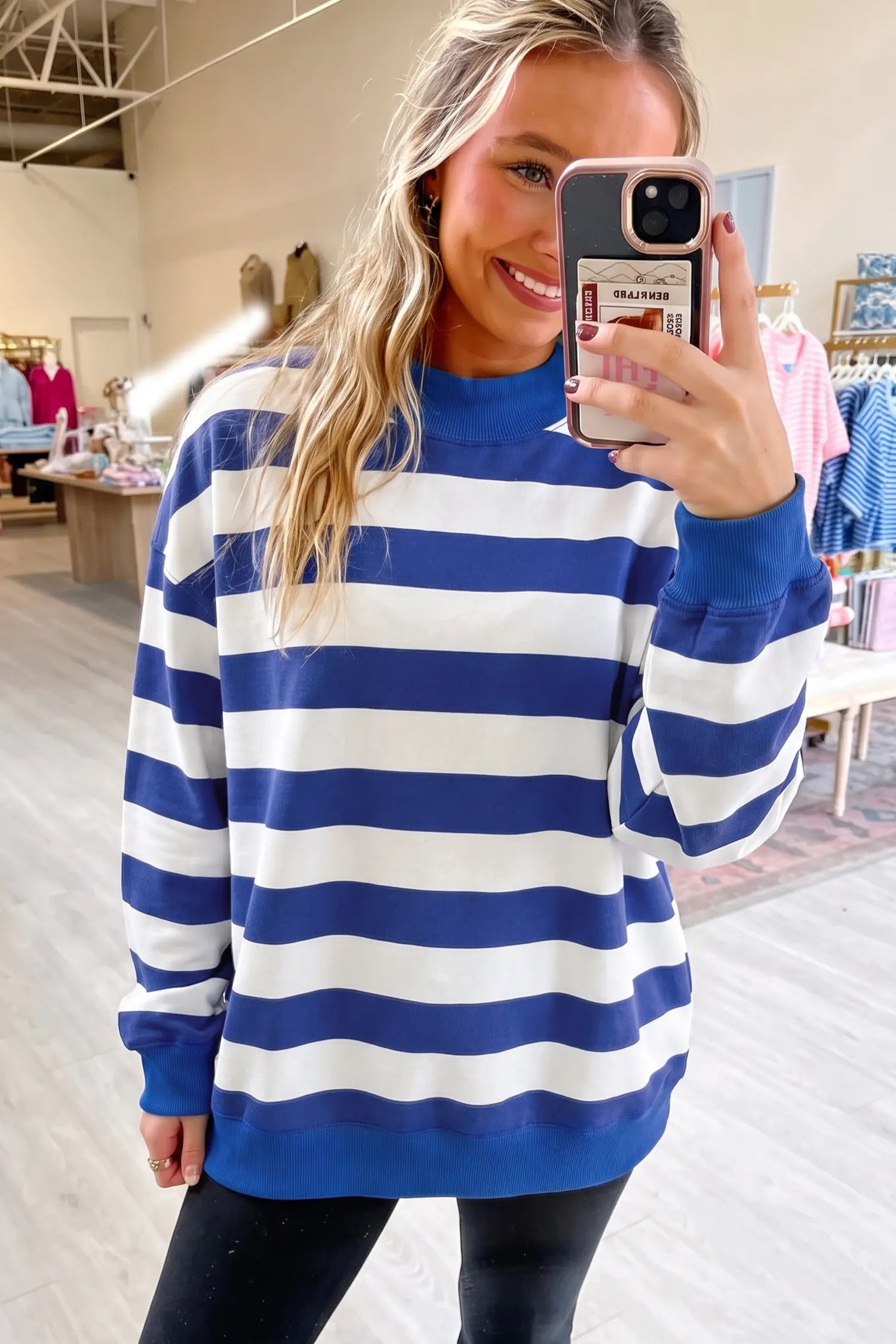 A woman taking a mirror selfie in a clothing store, wearing a blue and white horizontally striped crewneck sweatshirt.