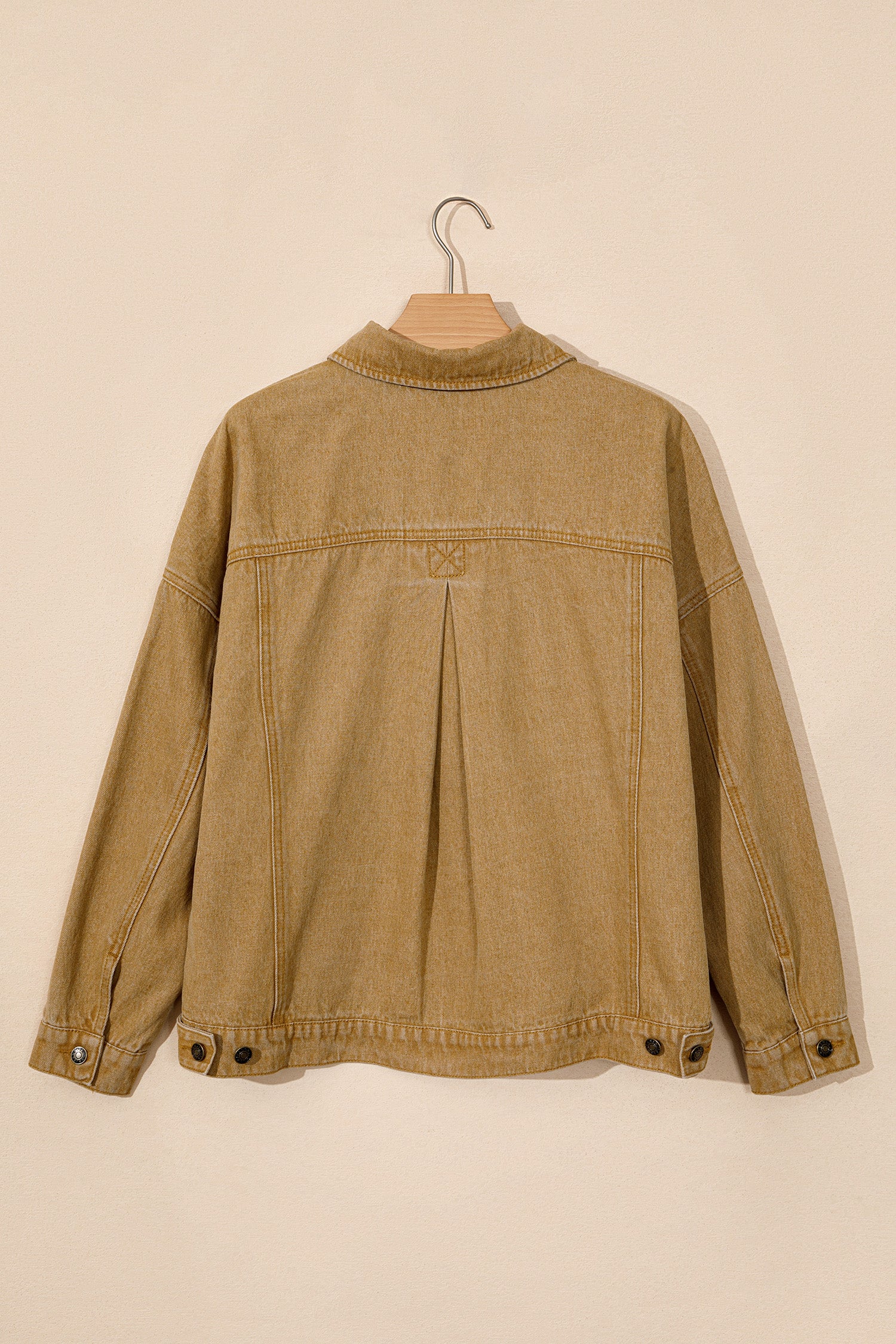 Tan denim jacket back view with pleats and button details, hanging on a wooden hanger against beige background.