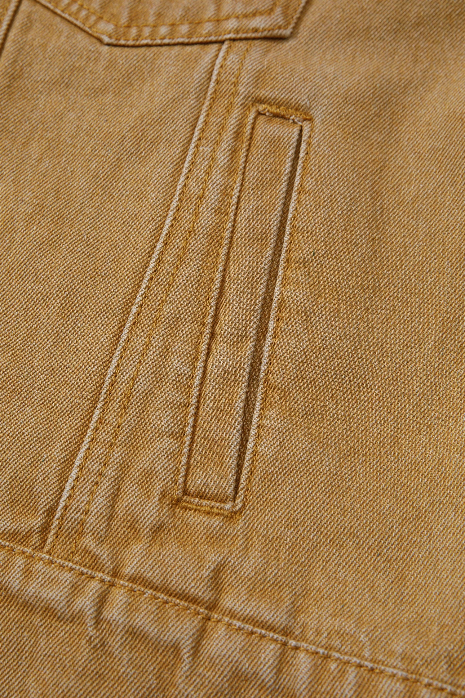 Close-up of a tan or camel-colored fabric garment, showing a diagonal welt pocket with contrast stitching.