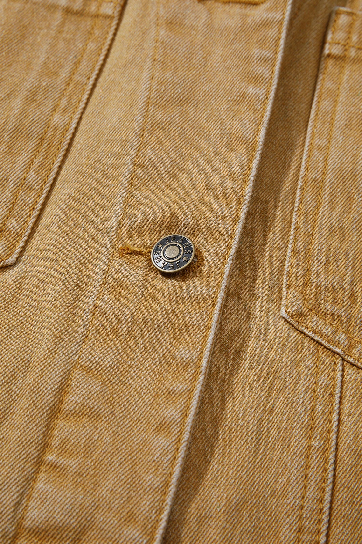 Close-up of a tan denim jacket showing the fabric texture, a button closure, and a side pocket.