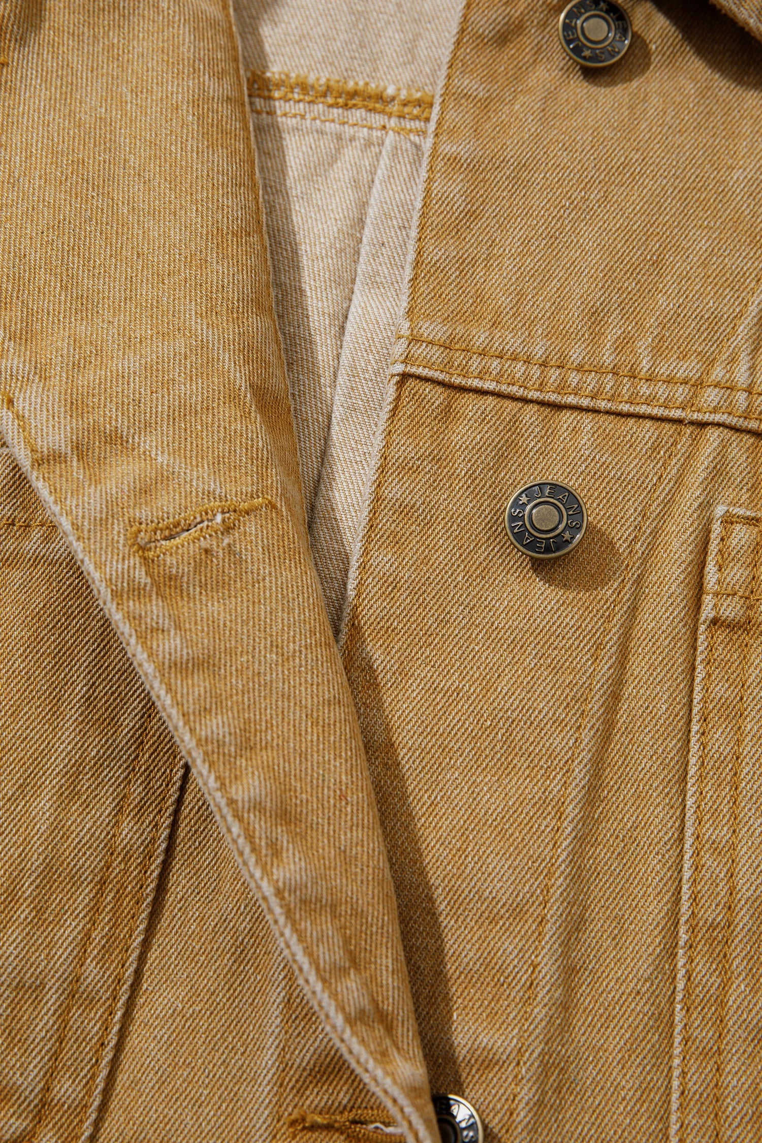 Close-up of a tan denim jacket showing the textured fabric, a silver metal snap button, and a light beige lining.