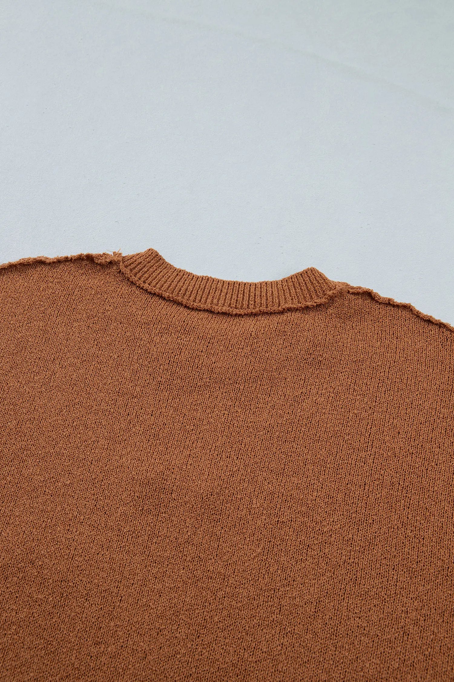 Close-up of a brown knit sweater showing the ribbed crew neck and textured fabric against a light gray background.