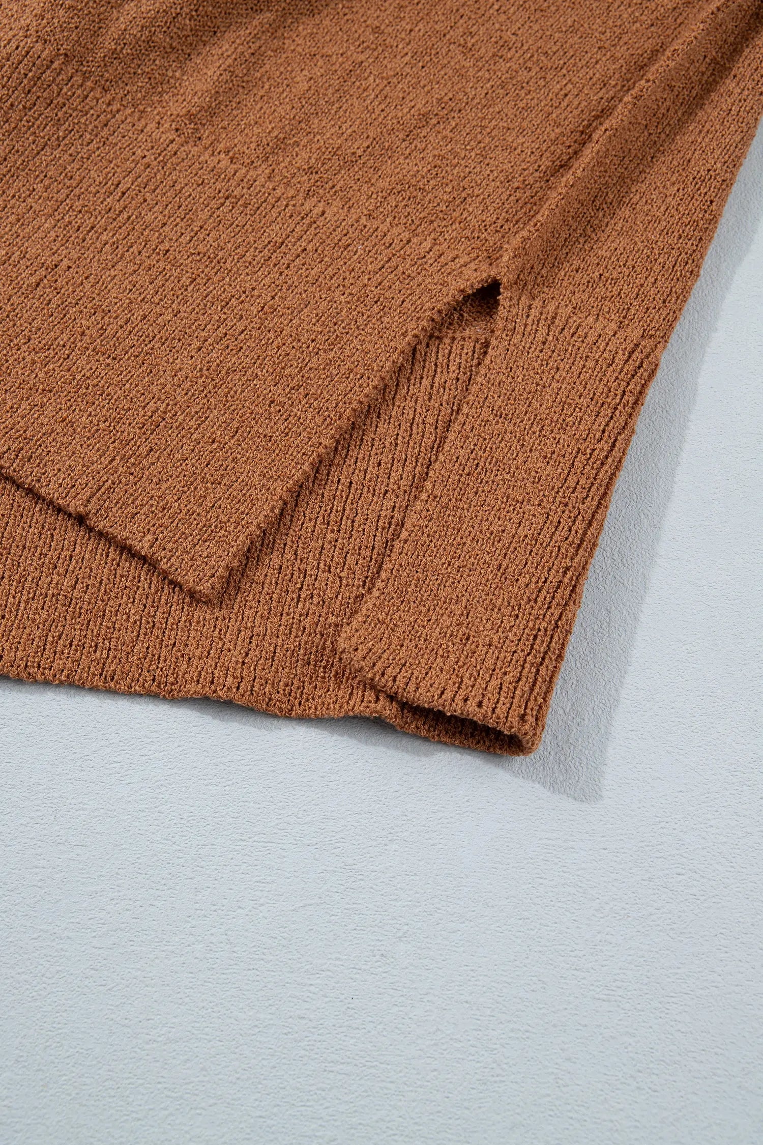 Close-up of a ribbed knit garment in a warm rust brown color, showing the texture and folded hem against a light gray background.
