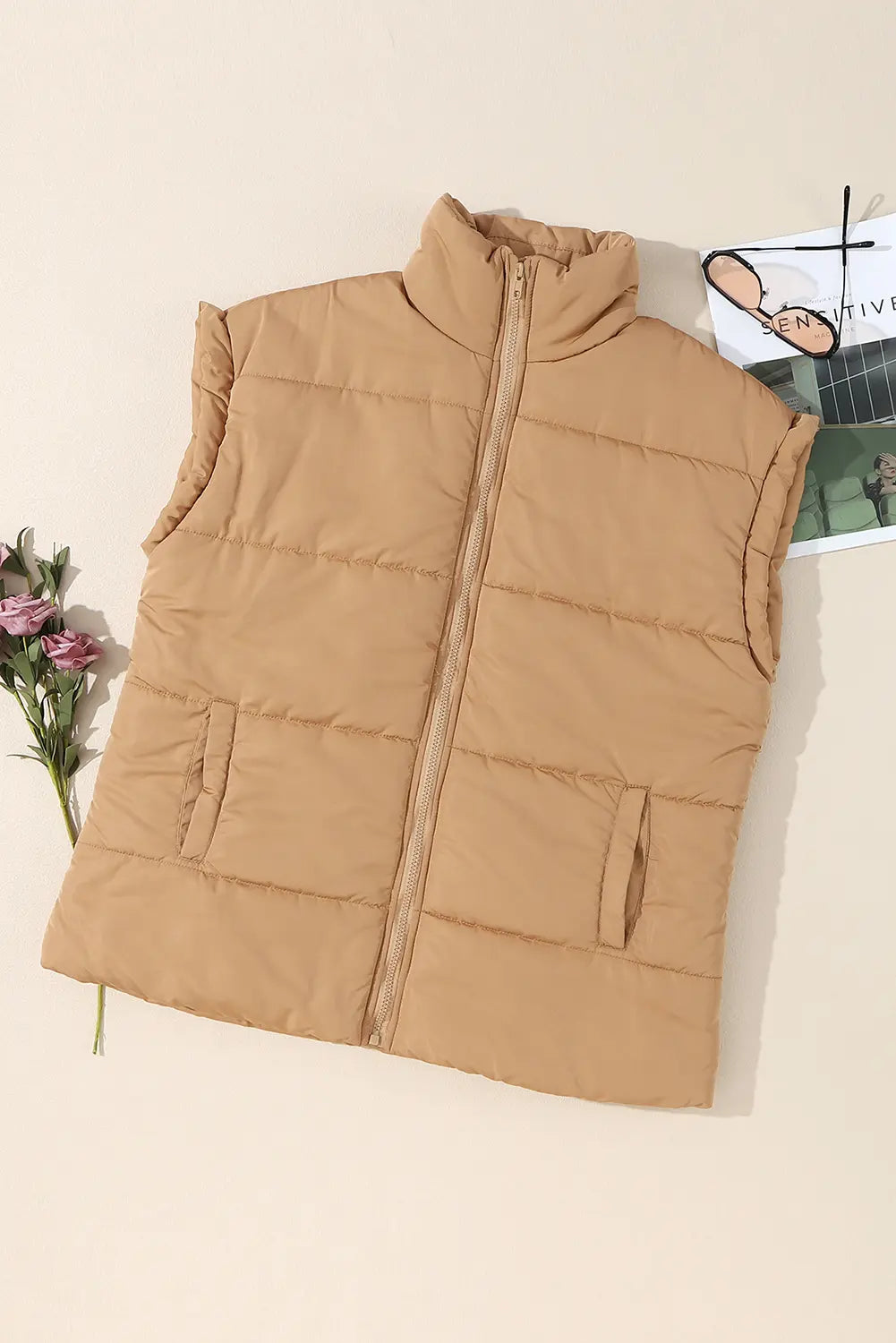 Woman wearing camel tan quilted puffer vest over pink knit sweater against a softly blurred outdoor background.
