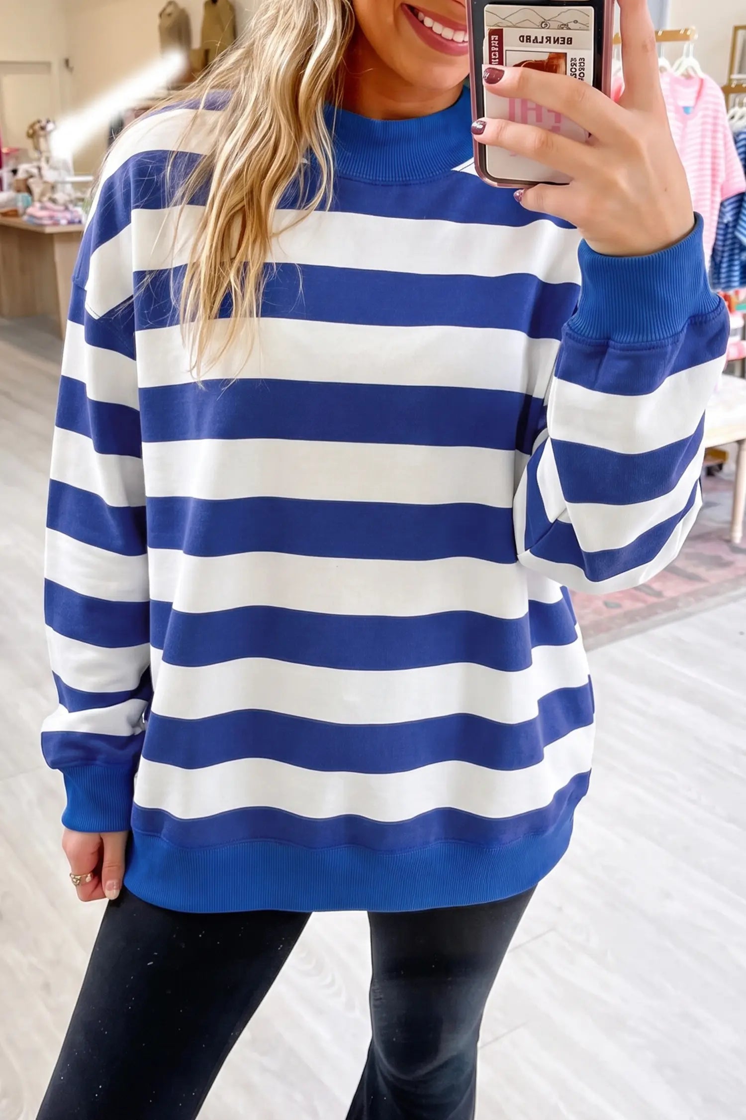 A woman takes a mirror selfie wearing a blue and white horizontally striped mock-neck sweatshirt and black pants in a retail store setting.