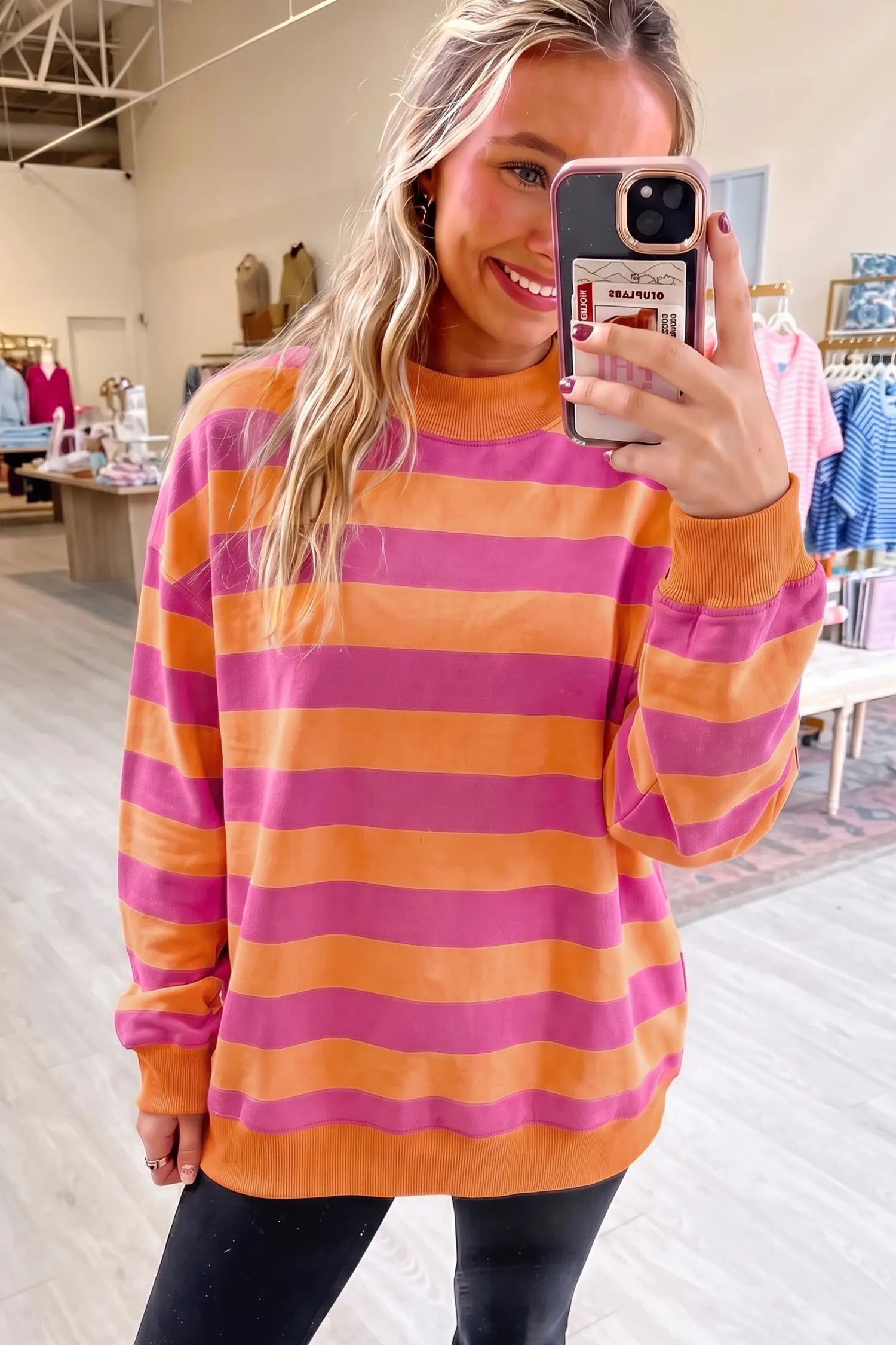 A woman takes a mirror selfie in a clothing store wearing a long-sleeve crewneck sweatshirt with horizontal stripes in orange and pink, paired with black pants.