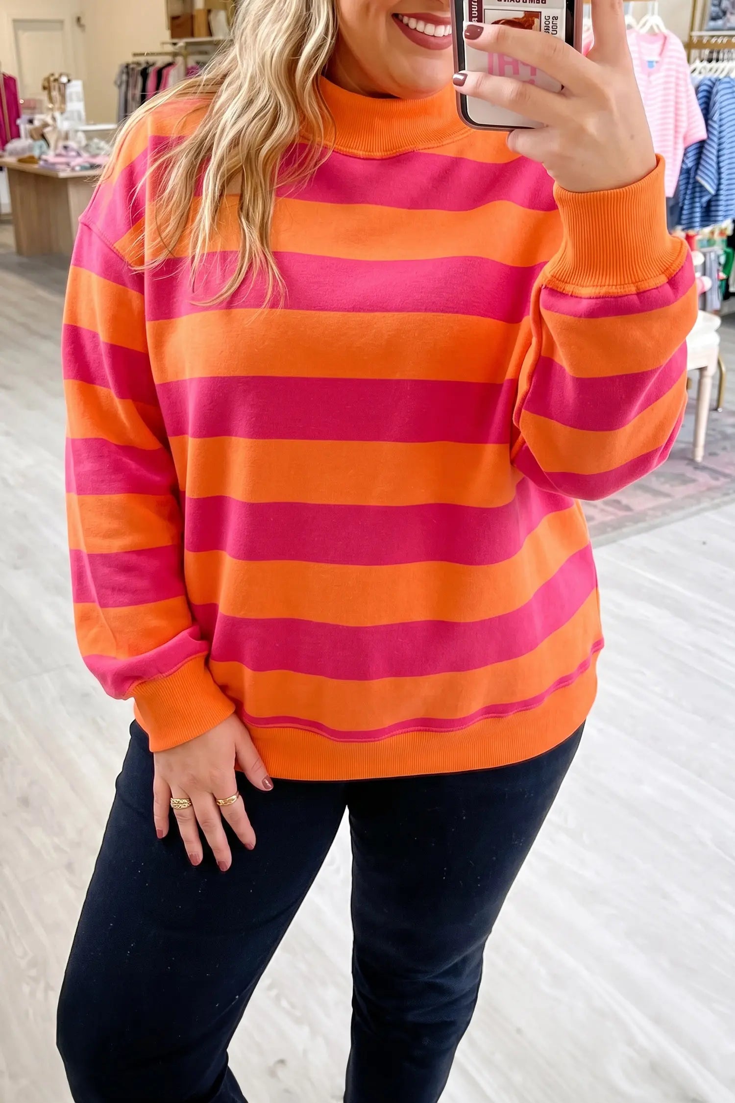 A woman takes a mirror selfie in a clothing store wearing a long-sleeve mock neck sweater with bold horizontal stripes in orange and pink, paired with dark jeans.
