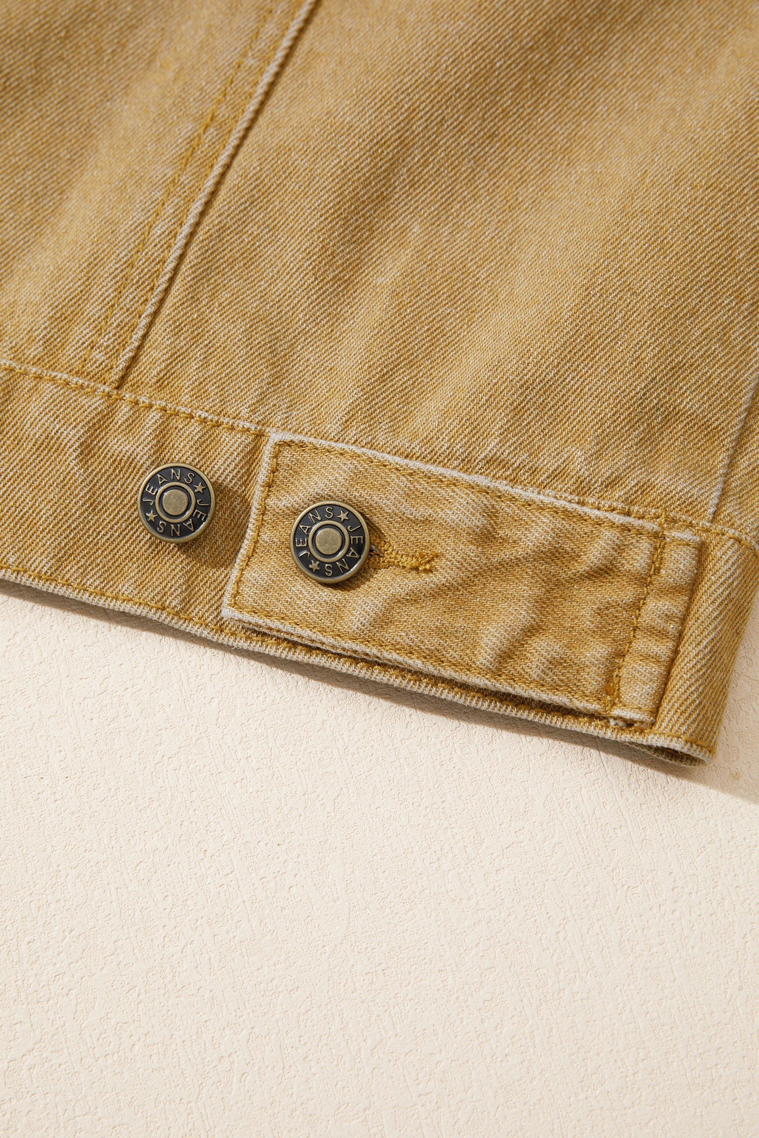 Close-up of a tan denim jacket showing the sleeve cuff with two dark metal buttons and a buttoned pocket flap.