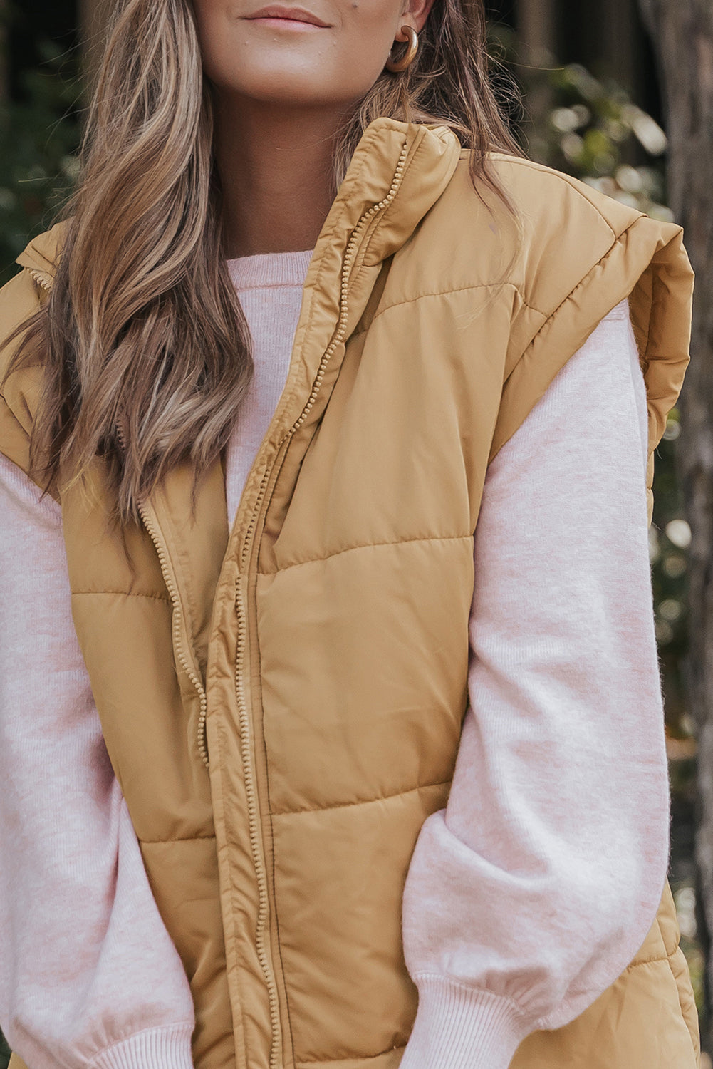 Woman wearing camel tan quilted puffer vest over pink knit sweater against a softly blurred outdoor background.
