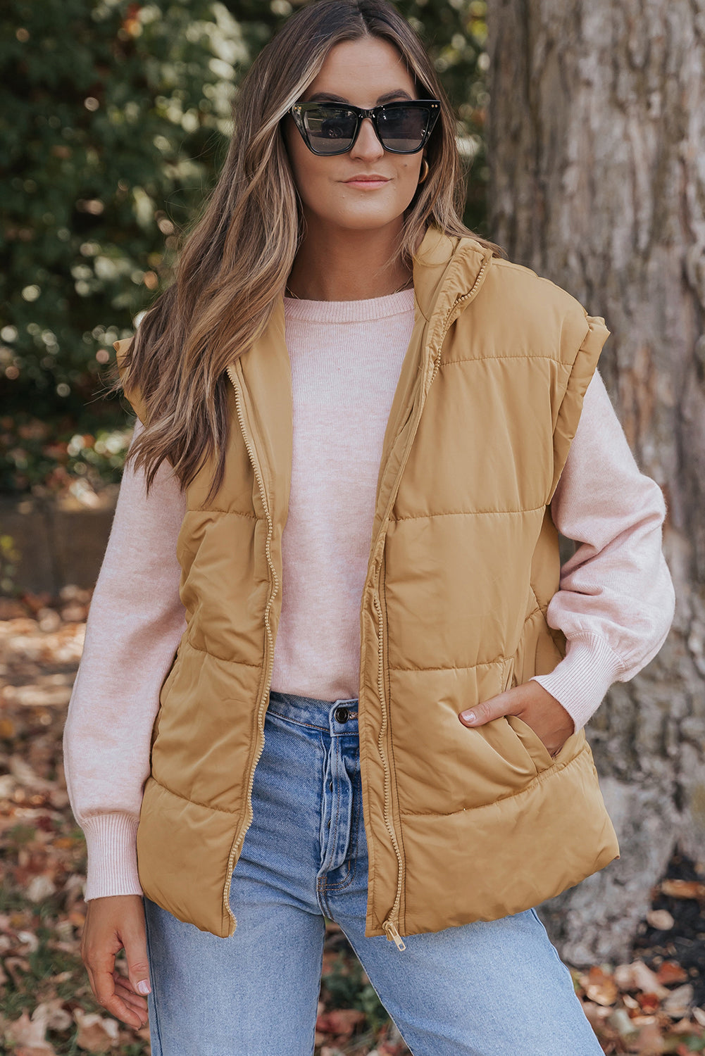 A woman wearing a tan quilted puffer vest over a light pink long-sleeve top and blue jeans, standing outdoors in front of a tree trunk.