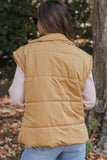 Woman viewed from behind wearing camel tan quilted puffer vest over pink shirt and blue jeans outdoors.