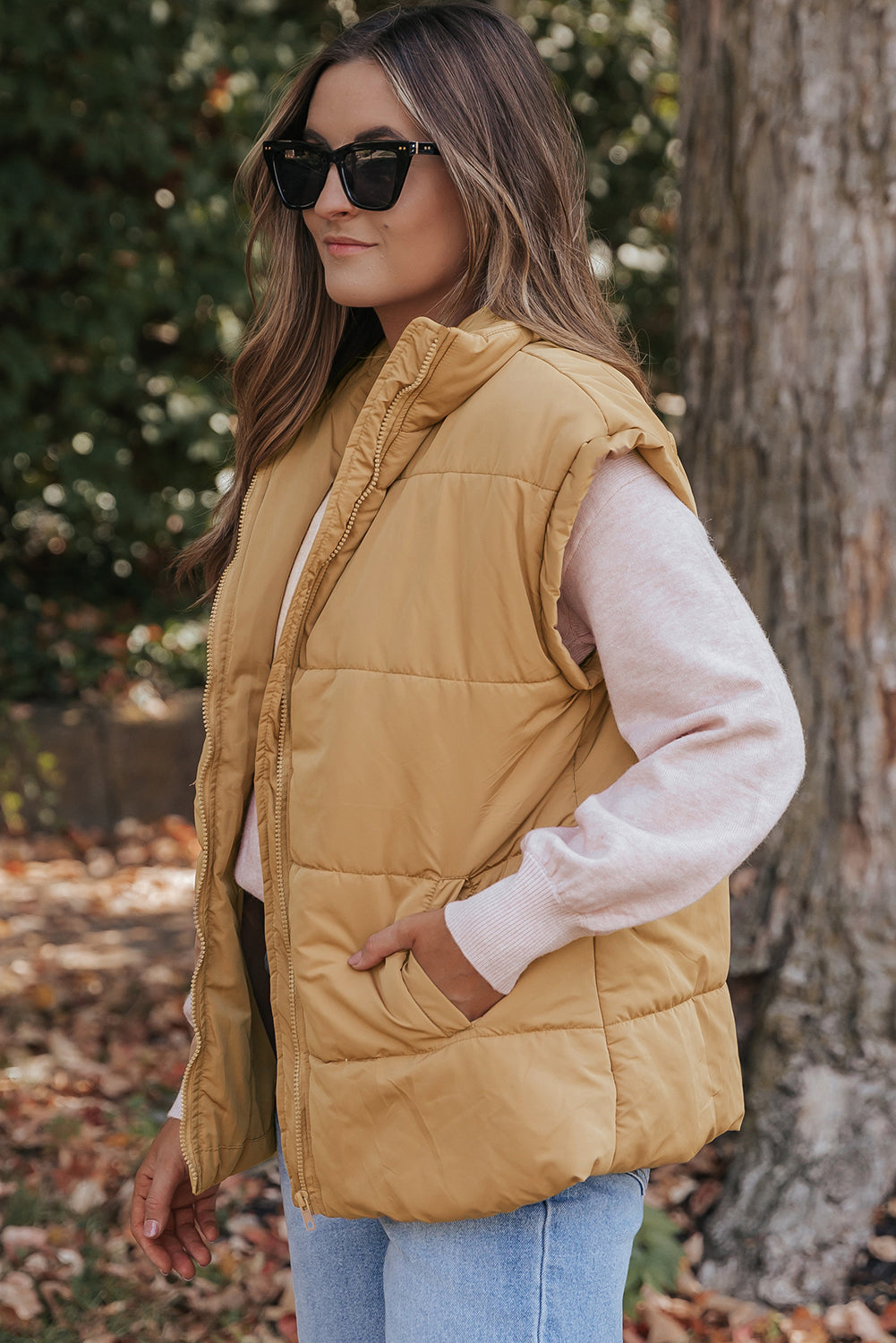 Woman wearing mustard yellow quilted puffer vest over pink top and blue jeans outdoors among fallen leaves.