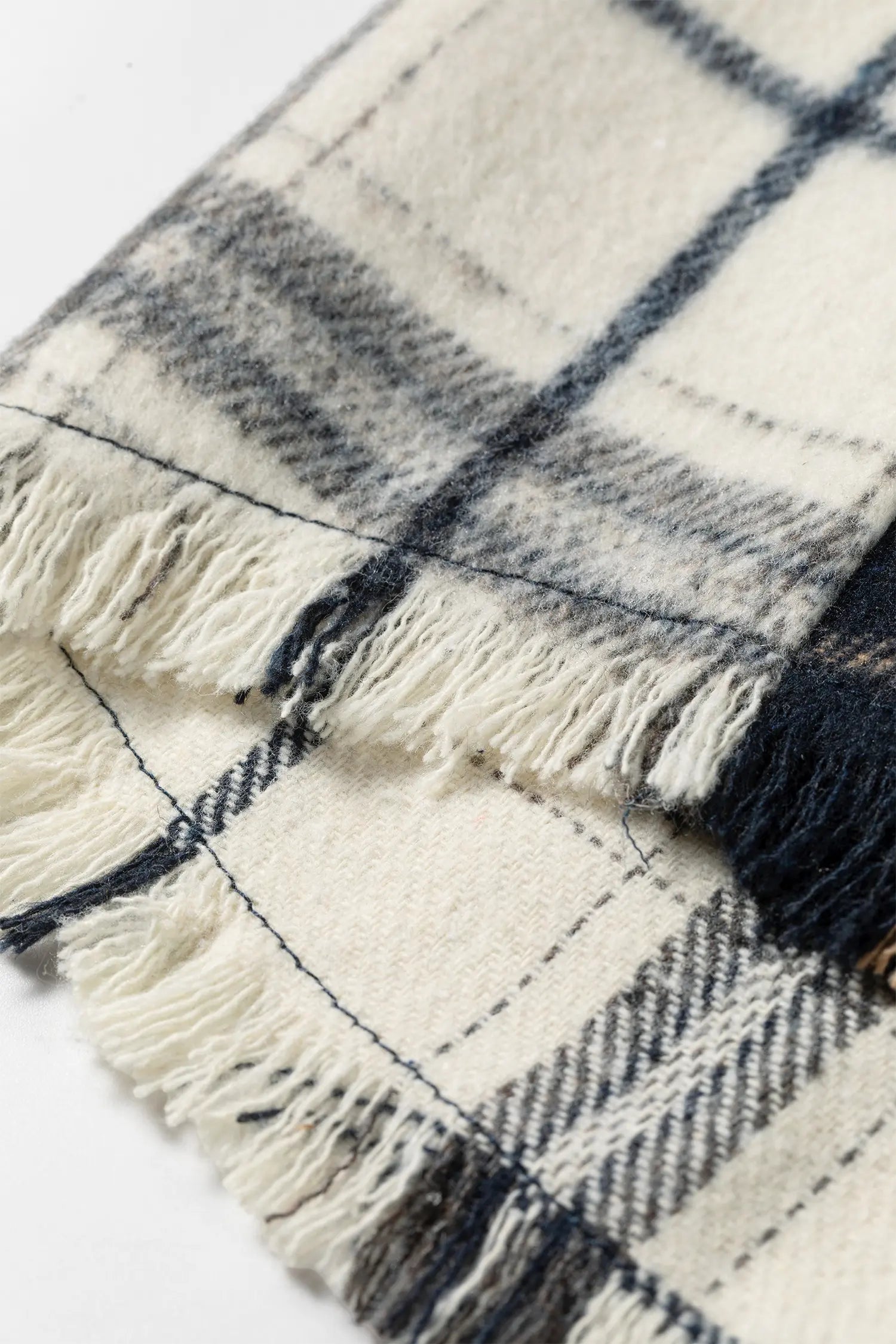 A close-up of a fringed plaid scarf or throw in off-white with dark navy and gray stripes, laid on a white background.