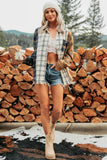 A woman stands outdoors in front of a large stack of cut firewood, wearing a cream knit beanie, a cream lace crop top, denim shorts, beige ankle boots, and a plaid button-up shirt layered over a tan jacket.