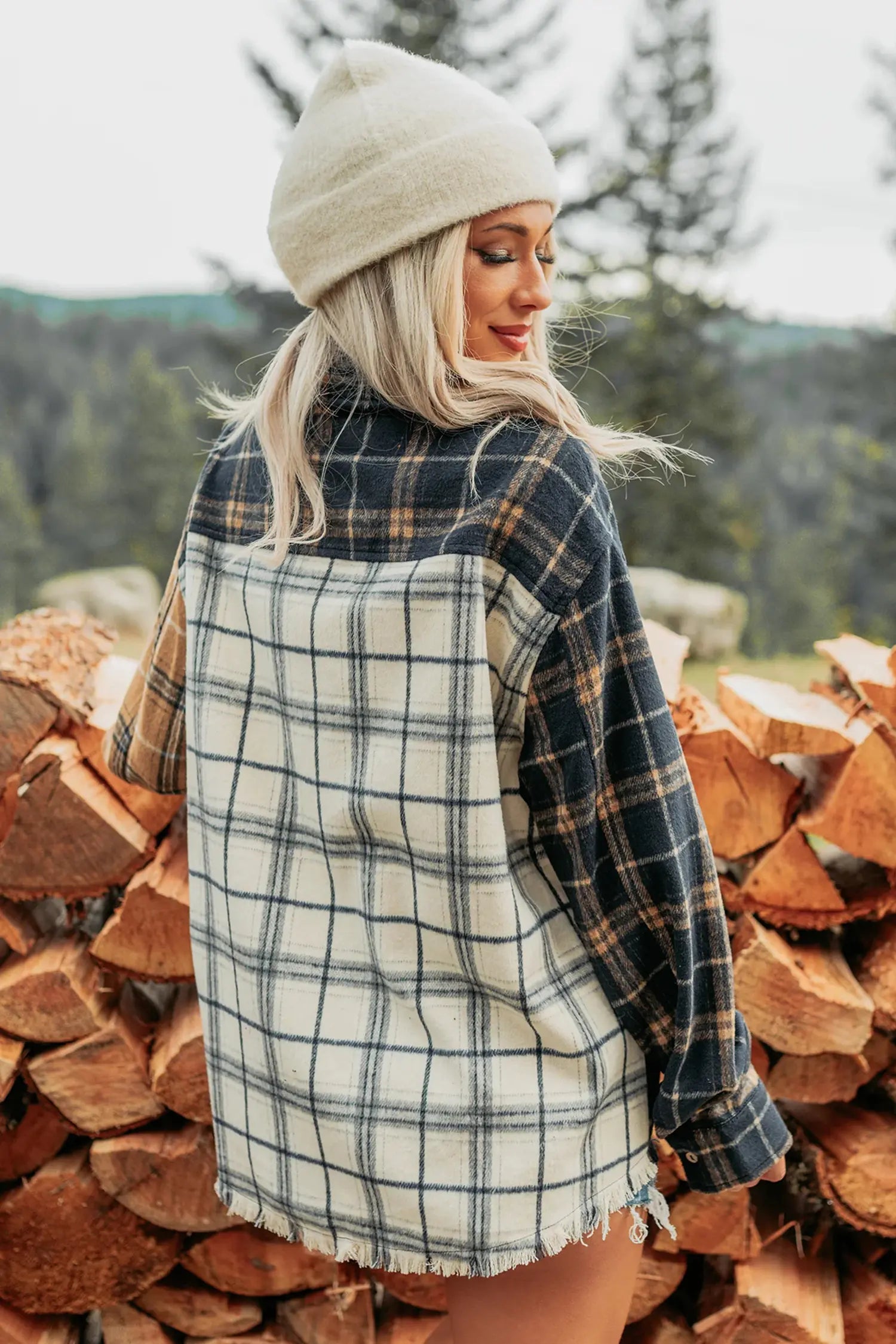 A woman wearing a patchwork plaid flannel shirt with navy, beige, and brown tones, a cream knit beanie, and standing outdoors in front of a stack of firewood.