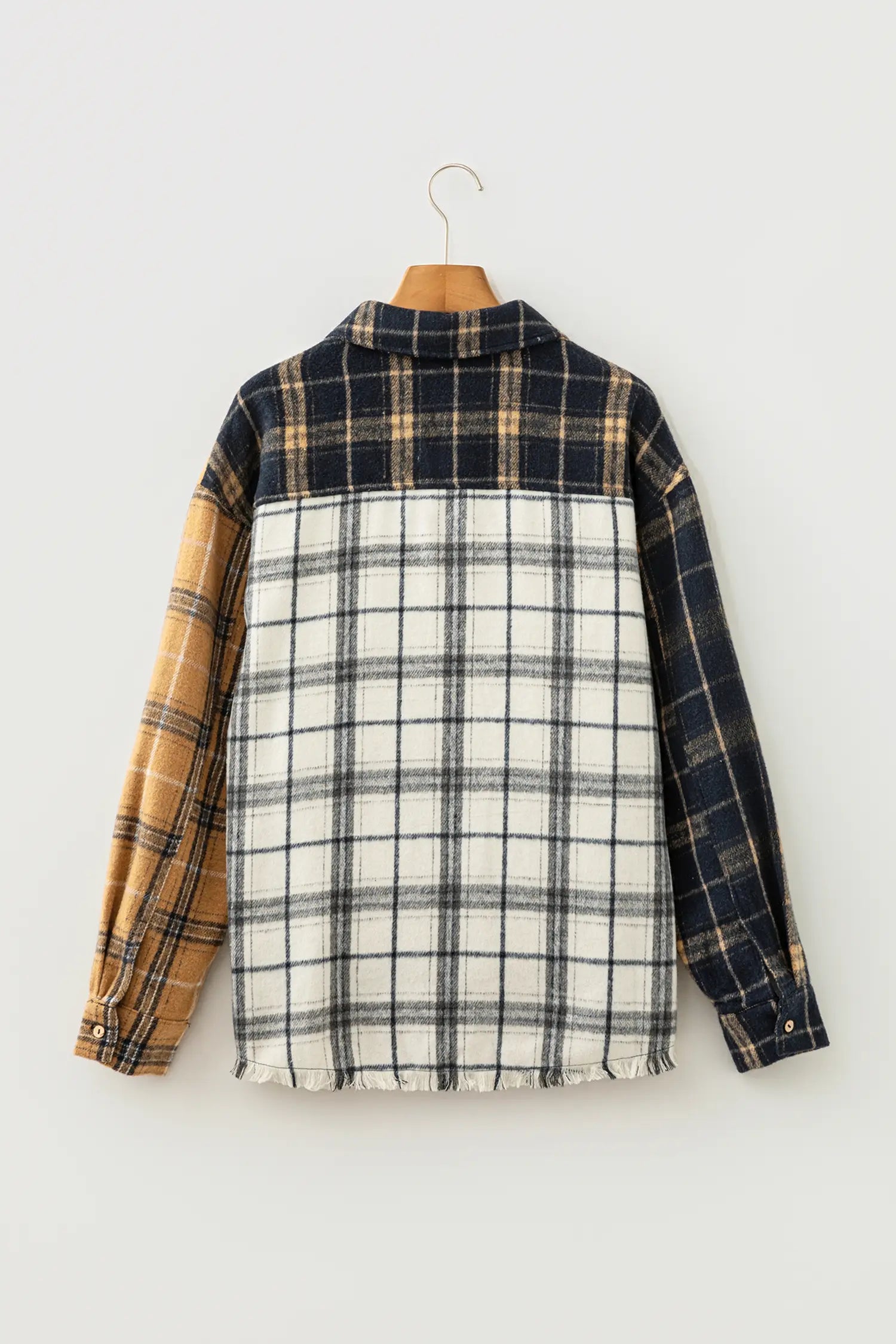 A long-sleeve plaid button-up shirt hanging on a wooden hanger against a plain light gray wall. The shirt features a navy and tan plaid yoke, a tan and cream plaid side panel, and a large cream, navy, and black plaid body.