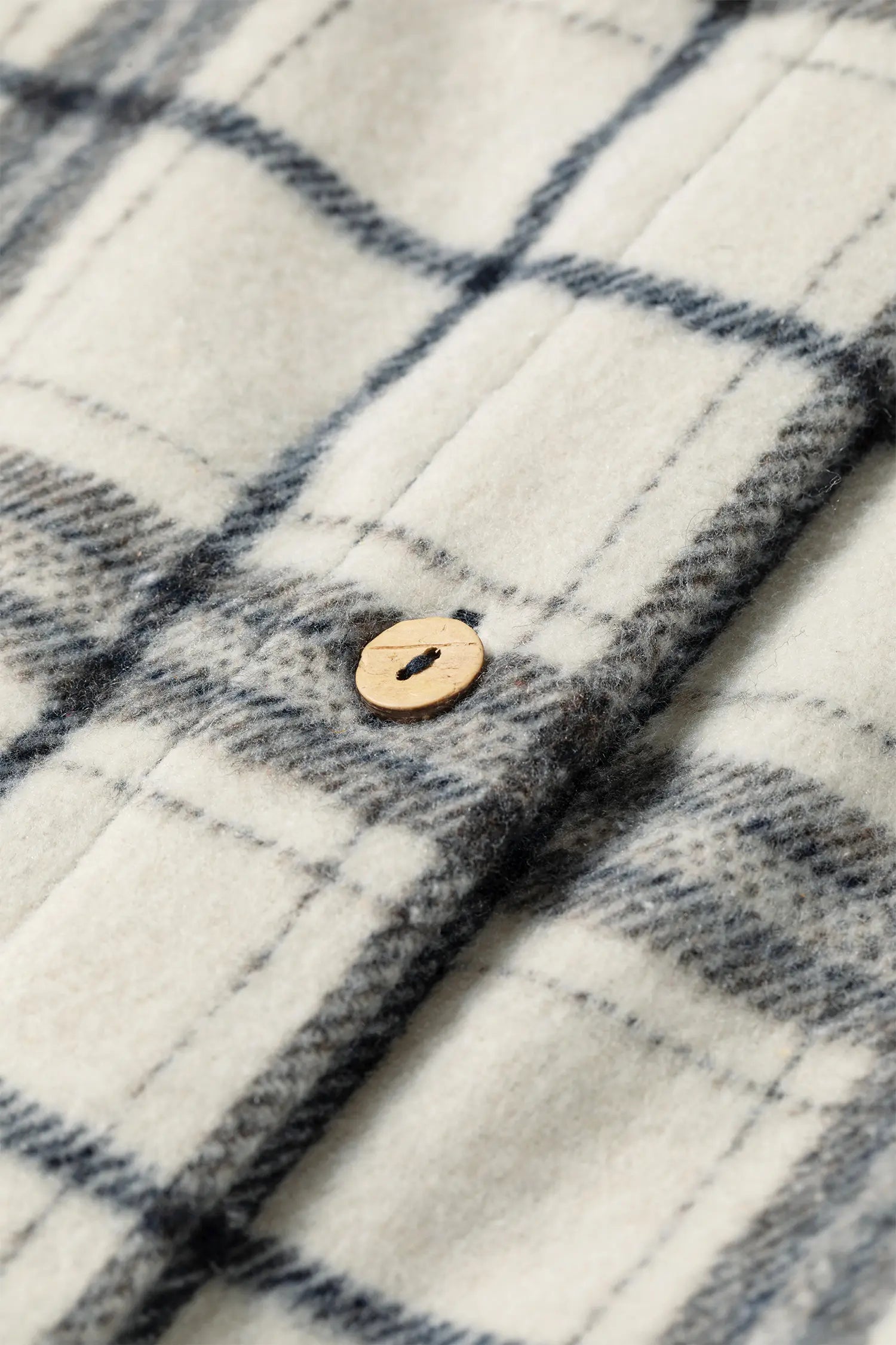 Close-up of a plaid fabric garment, likely a flannel shirt, in off-white with dark gray and navy lines, featuring a single light wood button.