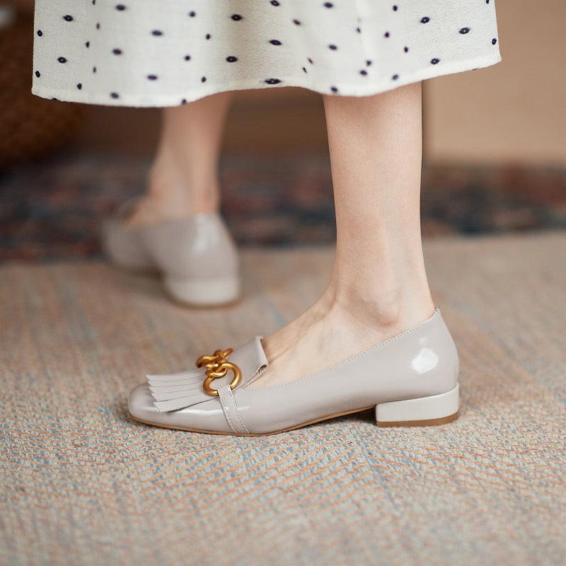 Light gray loafers with a gold chain detail worn by a person in a polka dot skirt.