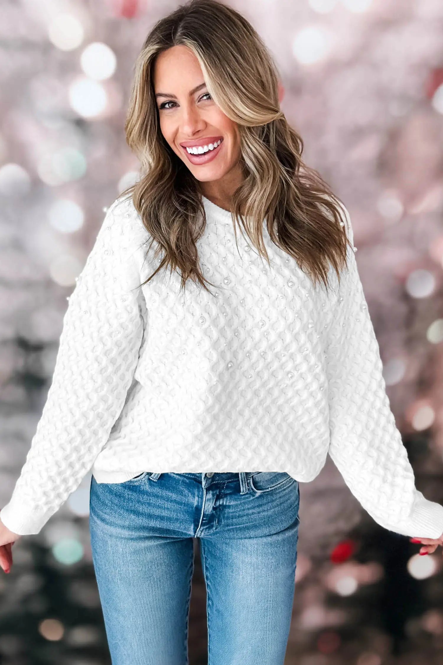 A smiling woman wearing a white textured knit sweater and blue jeans, standing against a soft pink bokeh background.