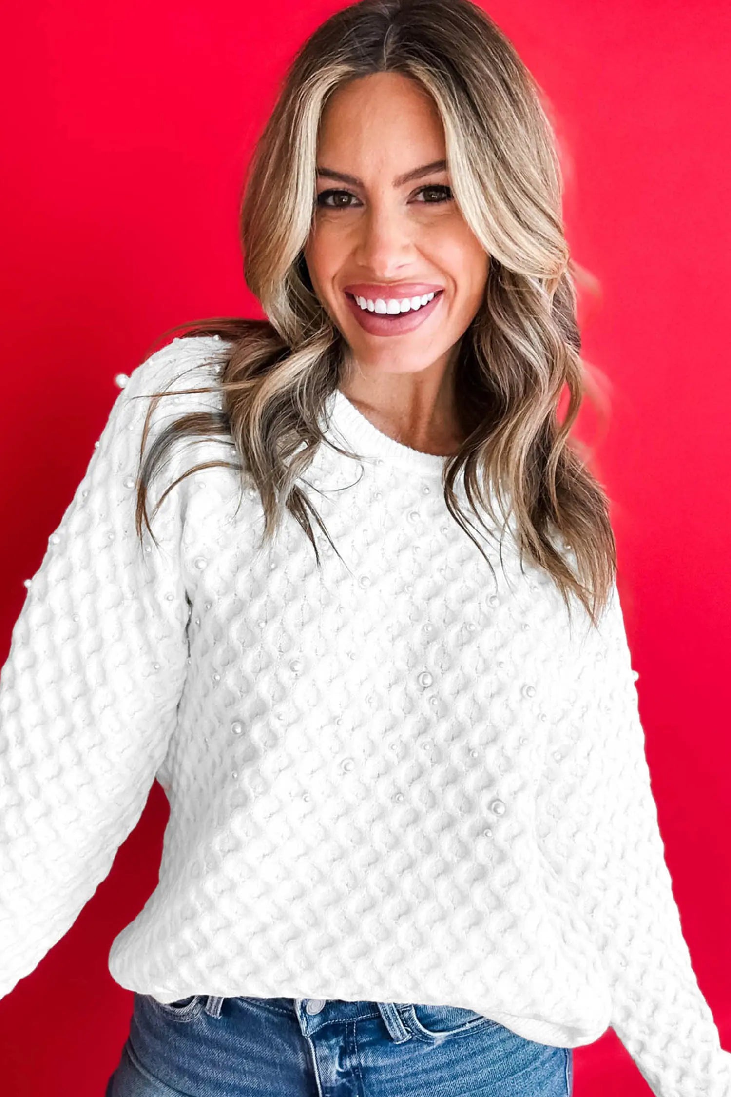 A smiling woman with blonde-highlighted hair wearing a white textured knit sweater with pearl-like embellishments and blue jeans, posed against a solid bright red background.