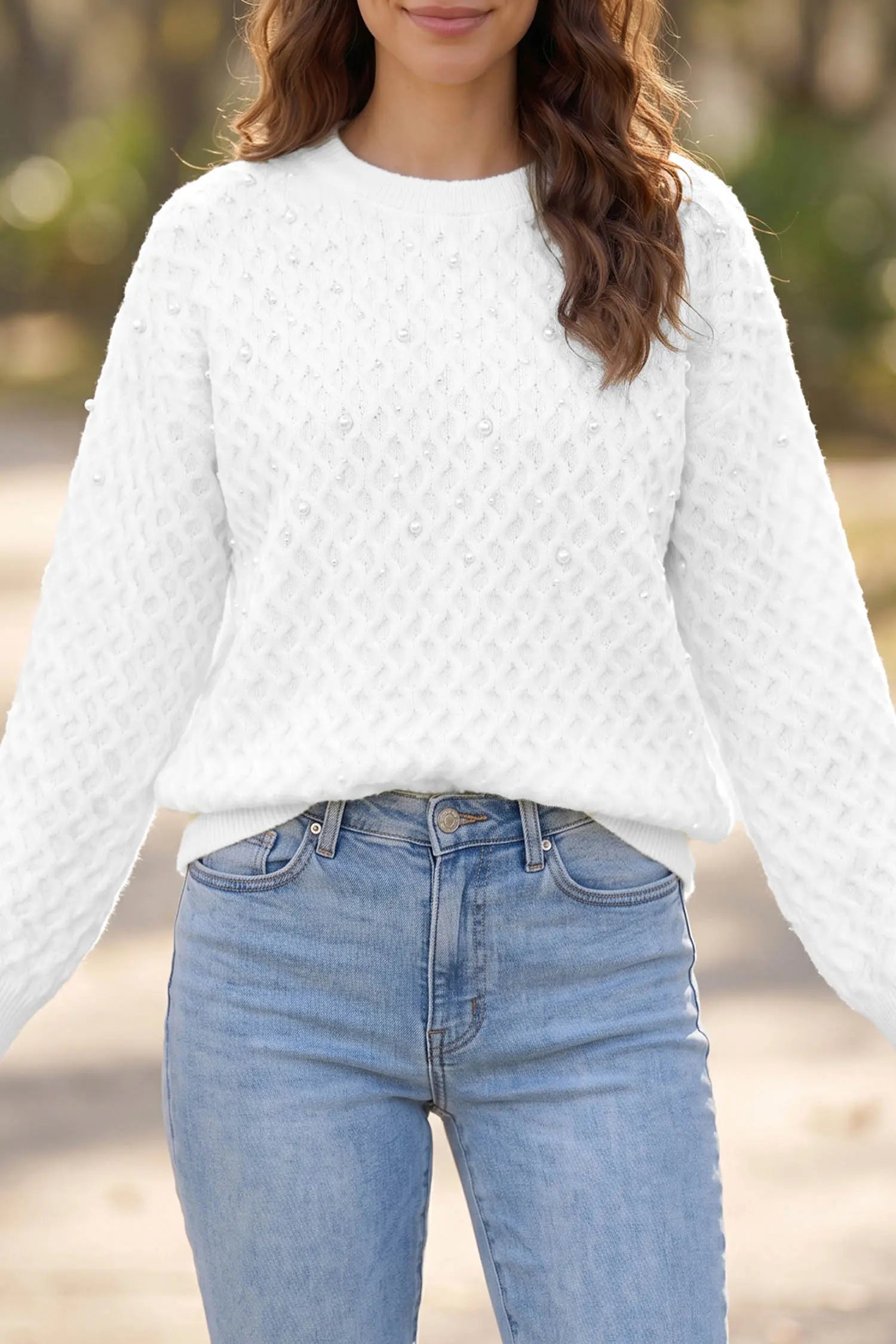 A woman wearing a white textured knit sweater with long sleeves and light blue jeans, photographed outdoors with a blurred background.