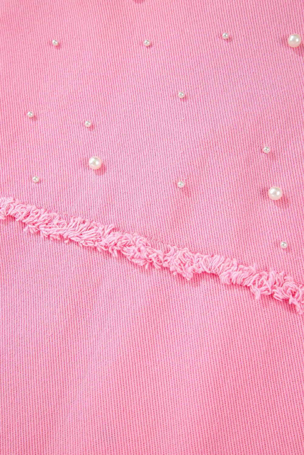 Close-up of a pink textured fabric with scattered small pearl-like embellishments and a frayed pink trim running horizontally across the middle.