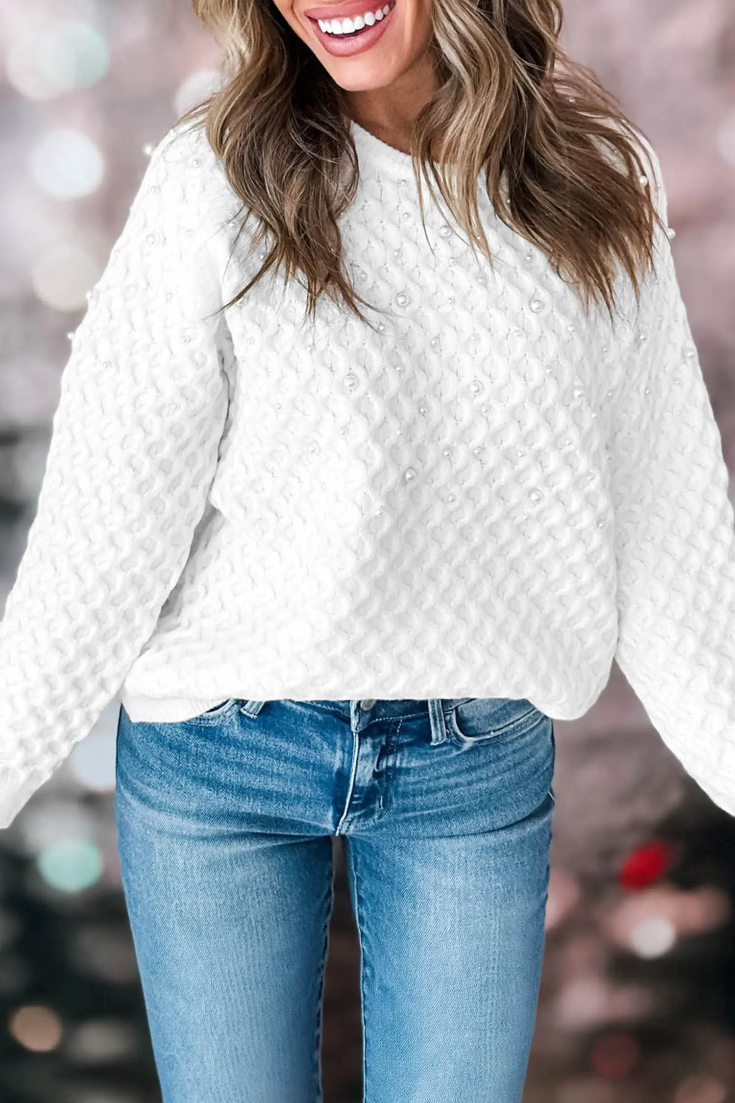 A woman wearing a white textured knit sweater with a diamond pattern and blue jeans, shown from the chest down against a blurred festive background.