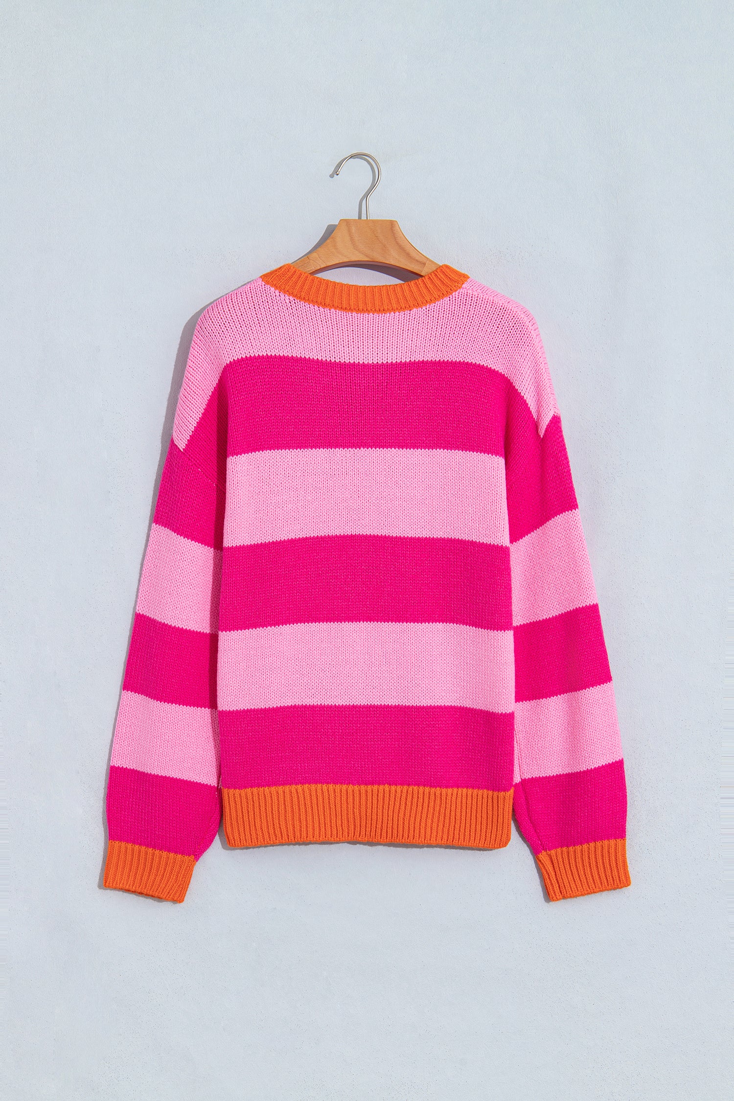 A pink and magenta striped knit sweater with orange ribbed trim, hanging on a wooden hanger against a light gray background.