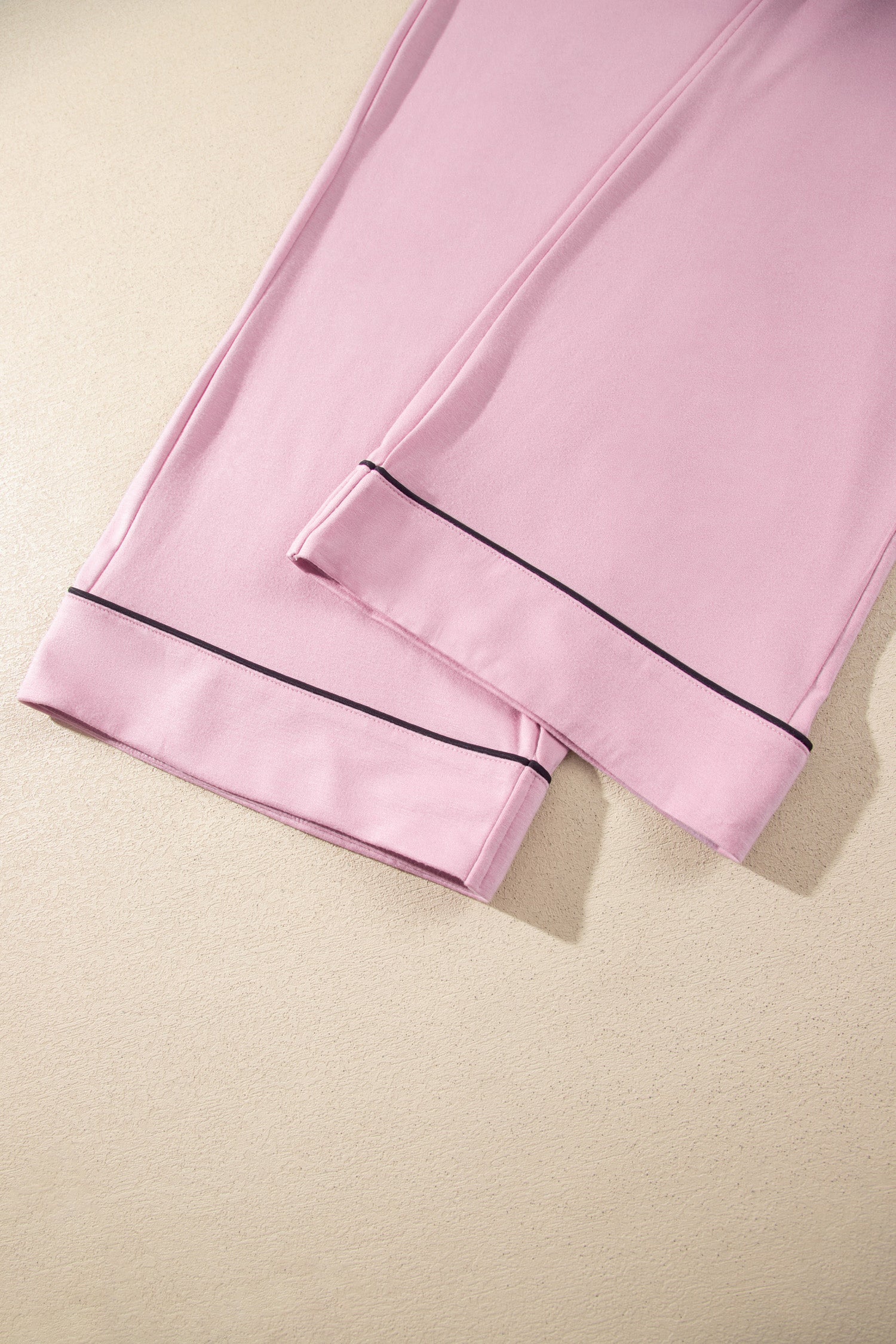 Pink pajama pants folded on a light beige textured surface, featuring thin black piping along the cuffs.