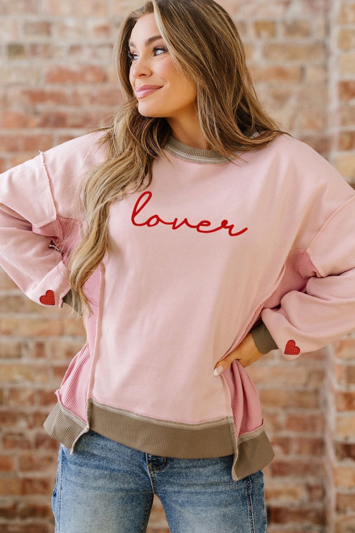 Woman wearing a pink sweatshirt with 'lover' text and heart designs, standing against a brick wall.