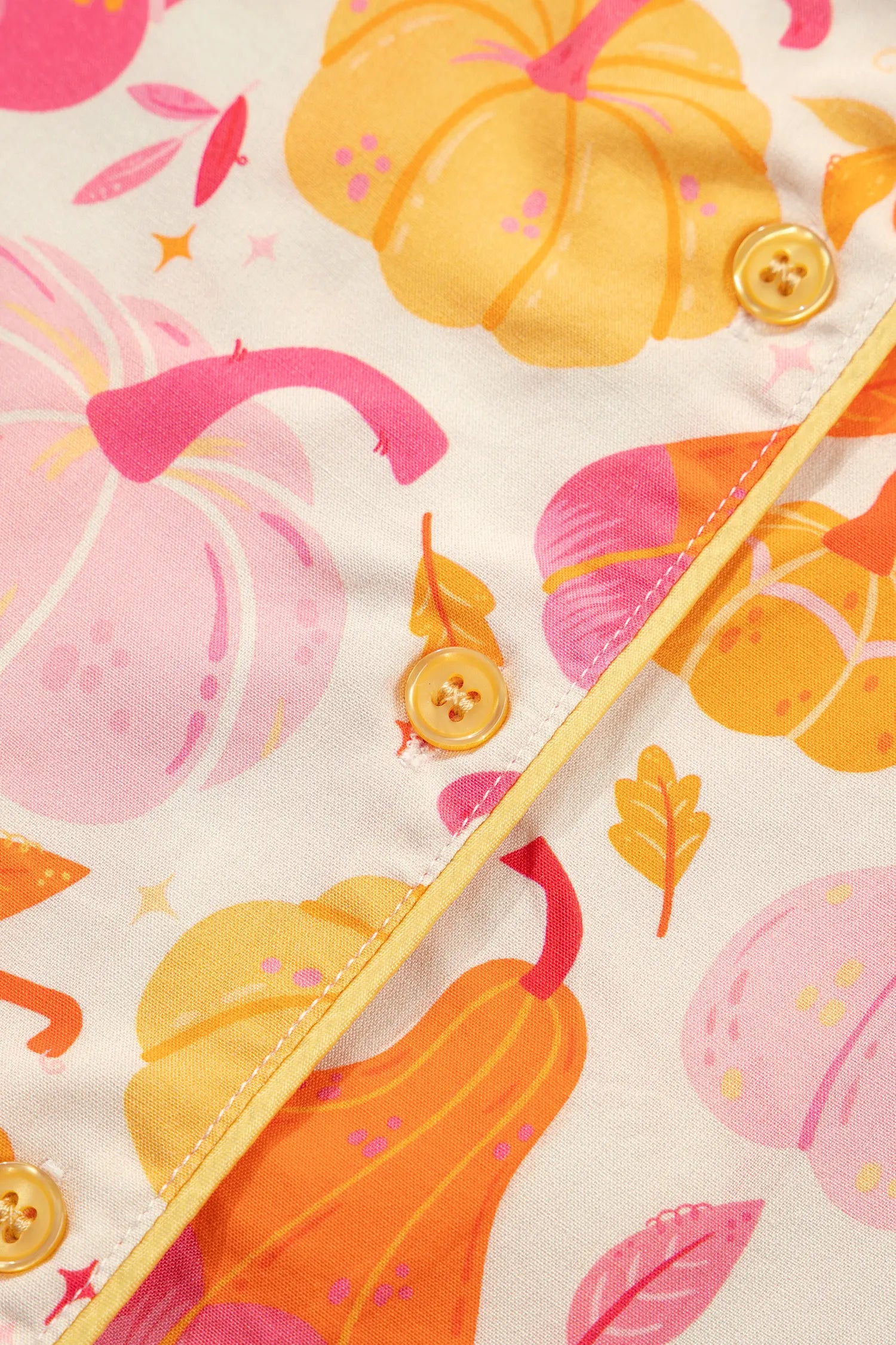 A close-up of a children's button-front garment featuring a colorful autumn print with pumpkins, gourds, leaves, and stars on a cream background. The fabric has a soft texture, and the edges show a light yellow trim with decorative buttons.