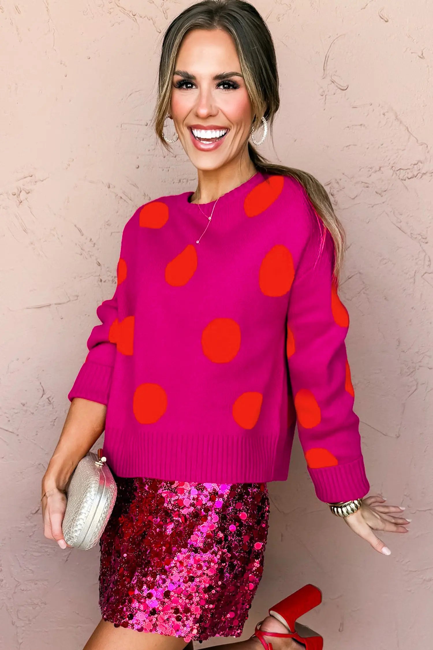 A woman modeling a bright pink crewneck sweater with large orange polka dots, paired with a shiny pink sequined mini skirt and holding a silver clutch against a textured pink wall.
