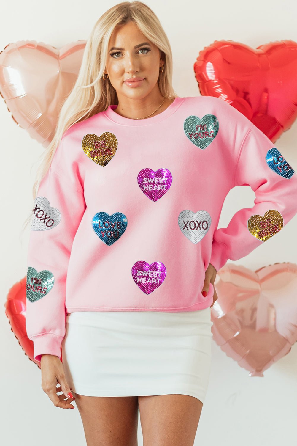 Woman wearing a pink sweatshirt with heart-shaped designs and text, surrounded by heart balloons.