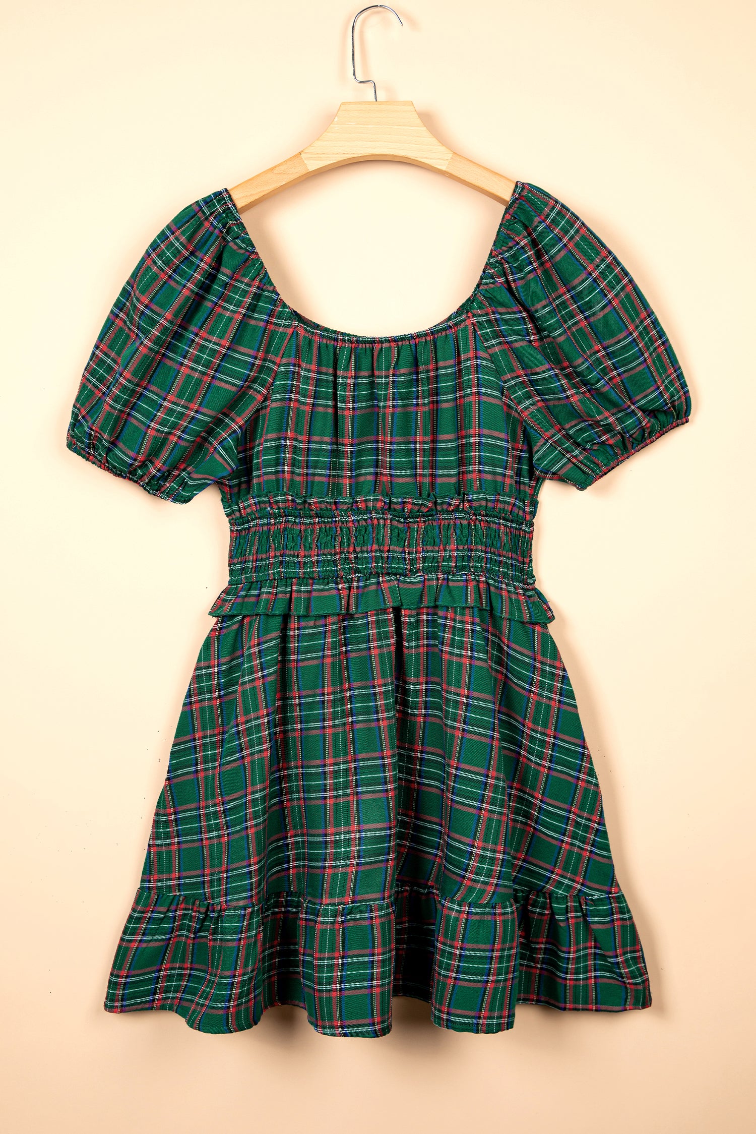 A green plaid short-sleeve dress with puff sleeves and a smocked waist, hanging on a wooden hanger against a solid beige background.