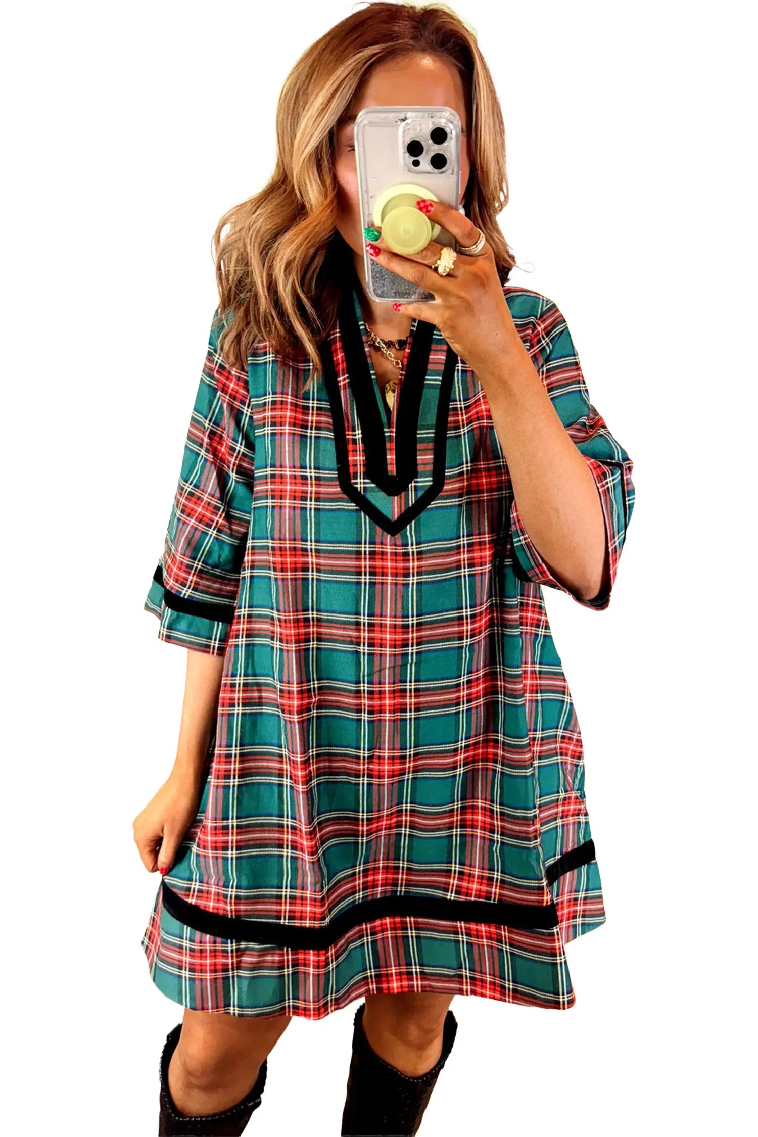 A woman takes a mirror selfie wearing a loose-fitting plaid tunic dress with three-quarter sleeves. The dress features a green, red, and white plaid pattern with black trim along the V-neckline and hem.