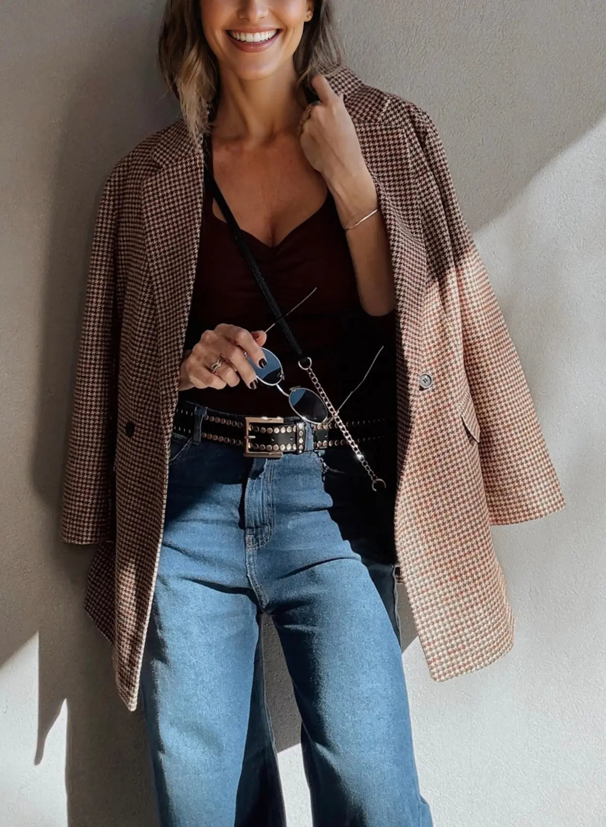 Woman wearing a plaid coat, black top, and blue jeans against a neutral wall.