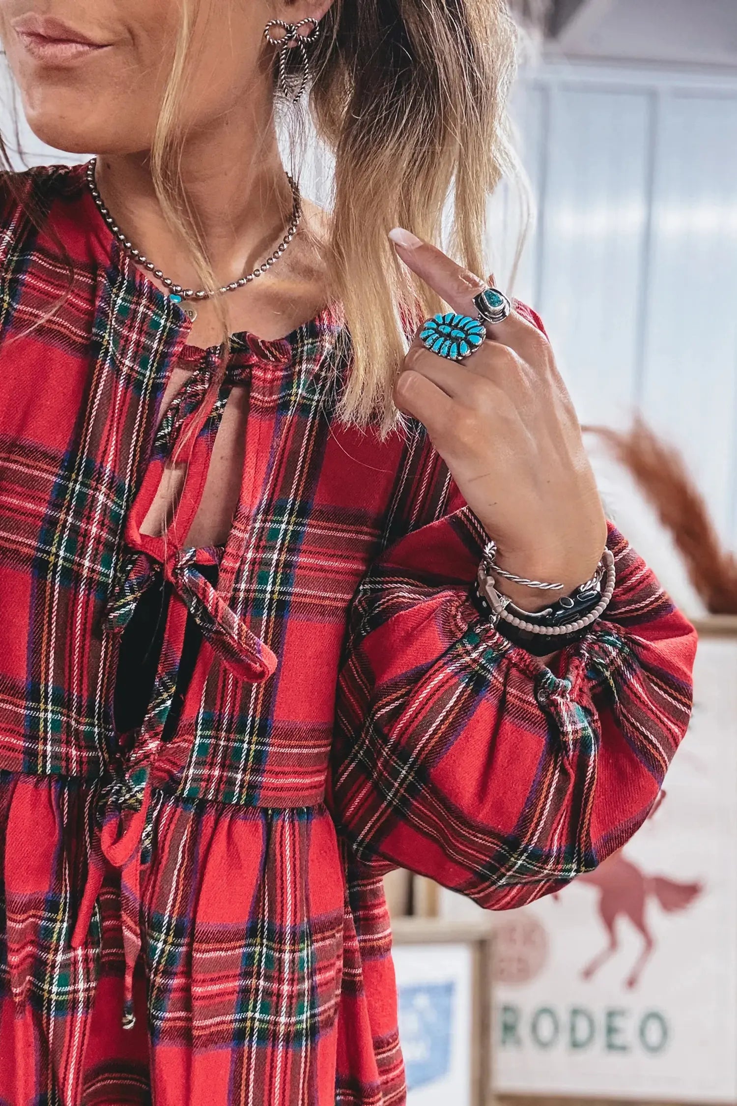 A close-up of a woman wearing a red plaid long-sleeve dress with tie-front details and puff sleeves, styled with layered necklaces and bracelets.