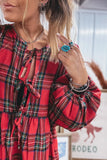 A close-up of a woman wearing a red plaid long-sleeve dress with tie-front details and puff sleeves, styled with layered necklaces and bracelets.