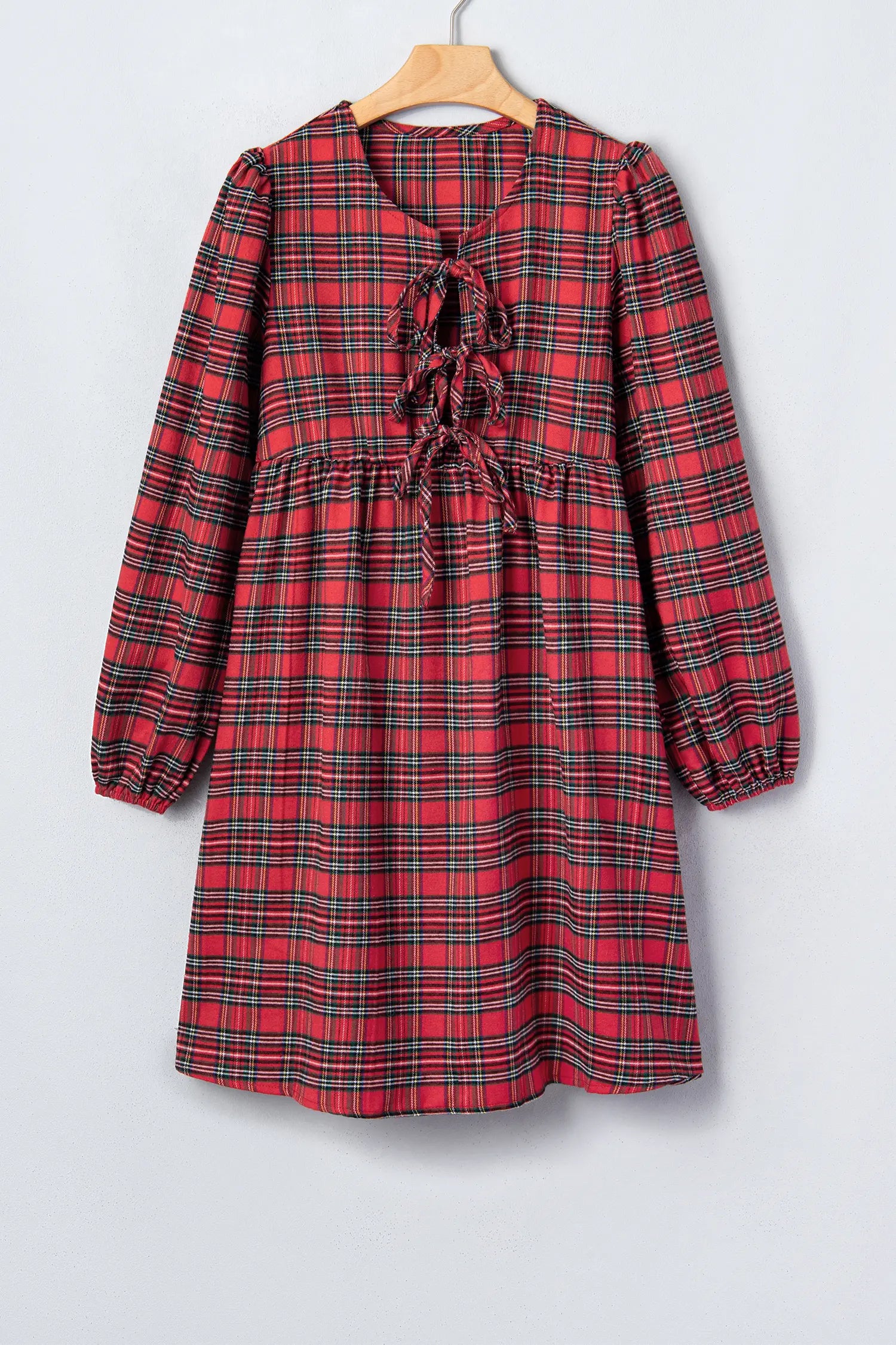 A red and black plaid long-sleeve dress with a ruffled tie neckline, hanging on a wooden hanger against a light gray background.