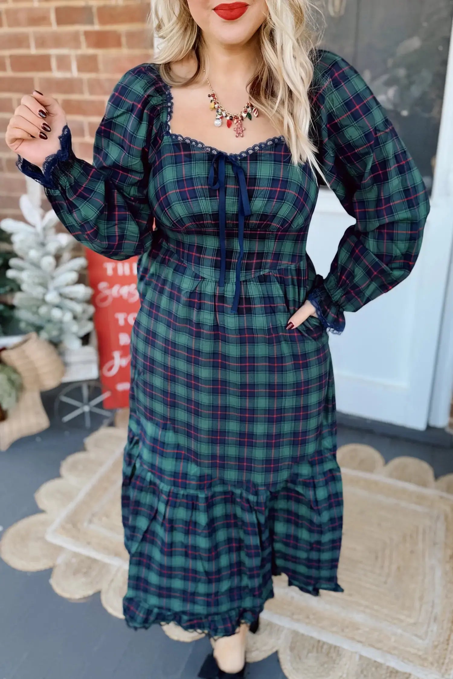 A woman wearing a long-sleeve plaid midi dress in dark green, navy, and red, standing on a porch with a brick wall and holiday decorations in the background.