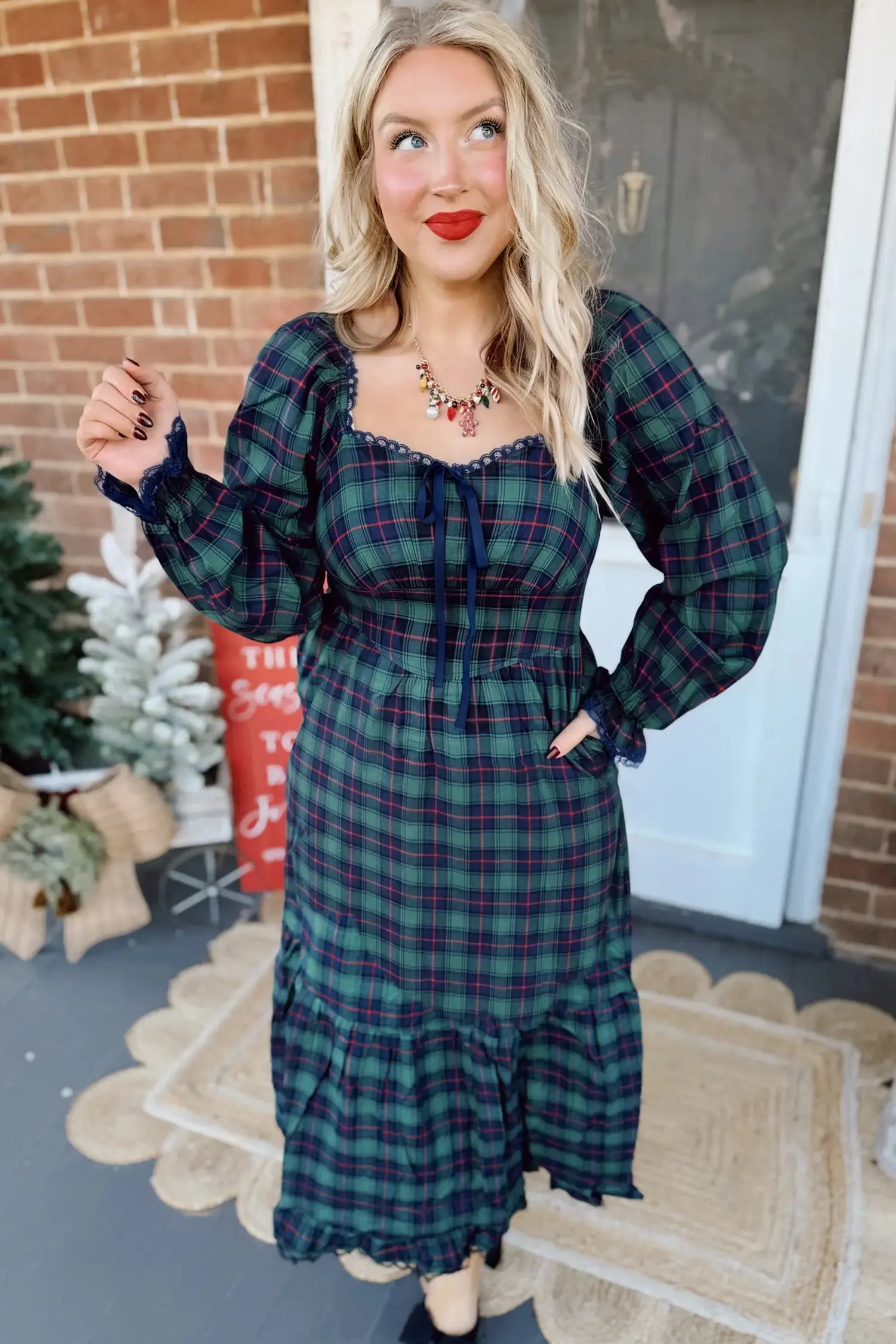 A woman wearing a long-sleeve plaid midi dress in dark green and navy, standing on a porch in front of a brick wall and white door.