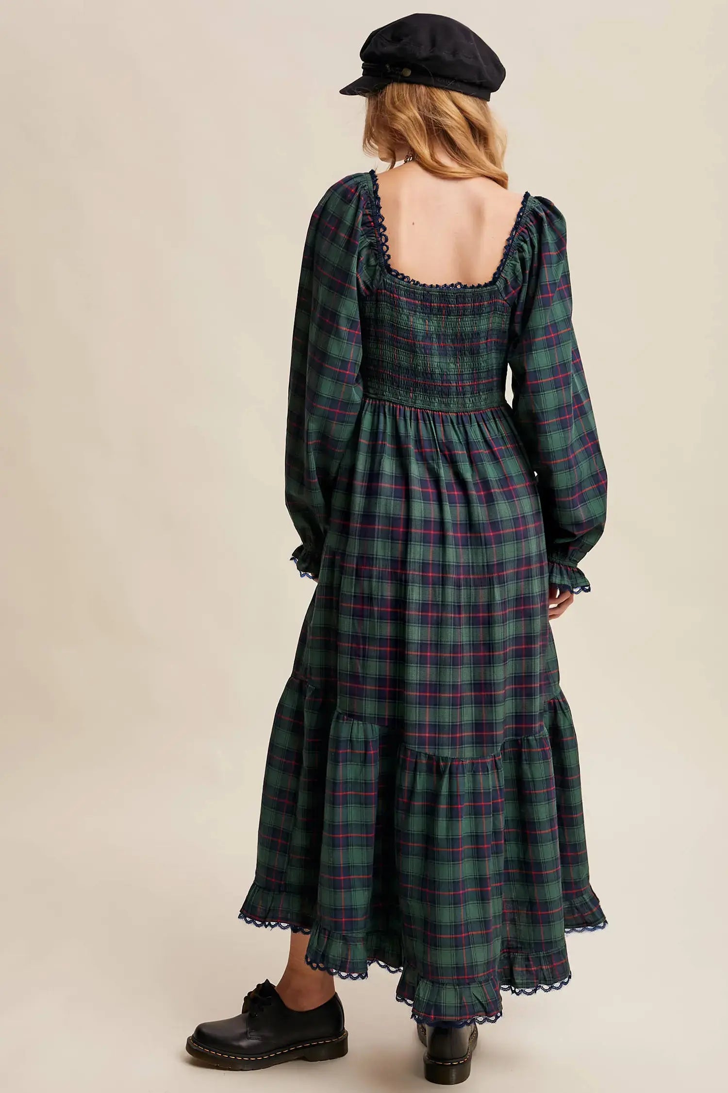 A woman shown from the back wearing a long-sleeved smocked maxi dress in a green and purple plaid pattern, paired with a black beret and black shoes, against a solid beige background.