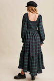 A woman shown from the back wearing a long-sleeved smocked maxi dress in a green and purple plaid pattern, paired with a black beret and black shoes, against a solid beige background.