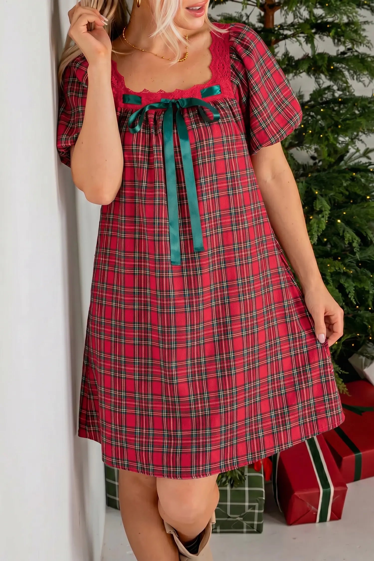 A woman wearing a red and green plaid short-sleeve dress with a teal bow at the neckline, standing indoors near a Christmas tree and wrapped gifts.