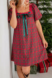 A woman wearing a red and green plaid short-sleeve dress with a teal bow at the neckline, standing indoors near a Christmas tree and wrapped gifts.
