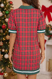 A woman wearing a red and green plaid short-sleeve dress with a green collar and white scalloped trim, photographed from the back in a holiday-themed room with a Christmas tree and fireplace in the background.