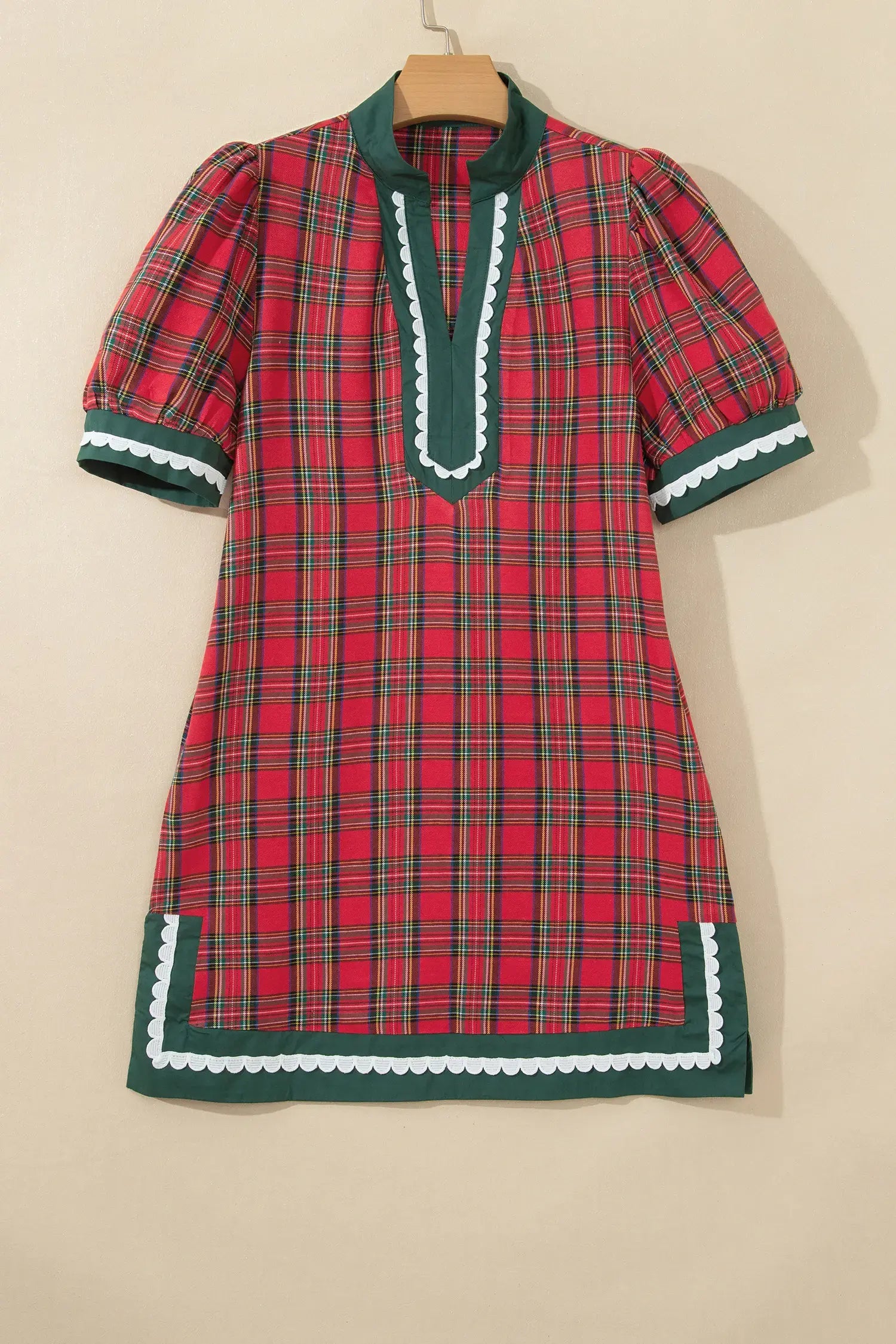 A short-sleeve plaid dress with puff sleeves, featuring a red and green tartan pattern with white scalloped trim details, hanging on a wooden hanger against a plain beige wall.