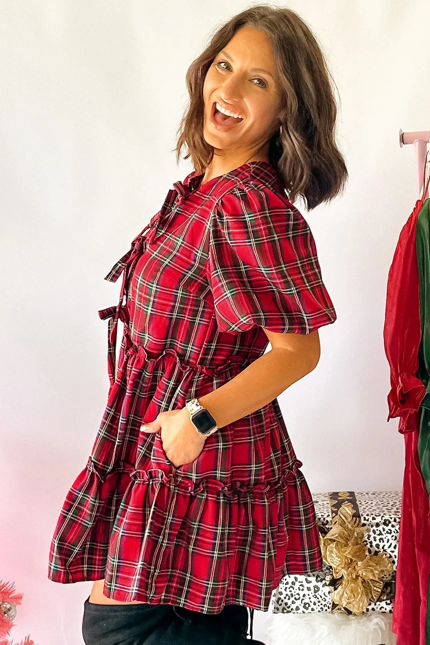 A woman modeling a red and black plaid short-sleeve tunic dress with tie details on the chest and a gathered waist, standing against a light pink wall.