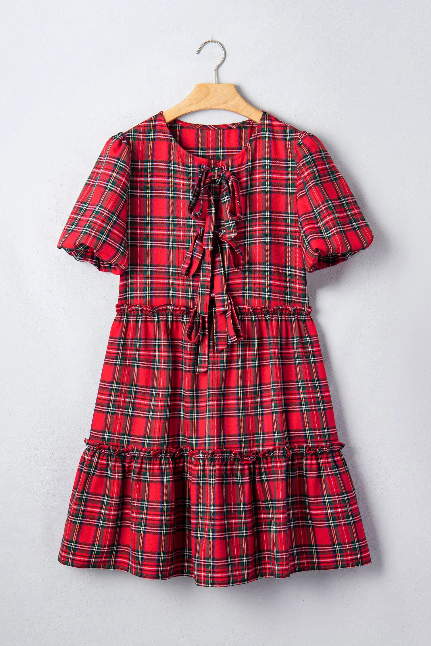 A red and green plaid short-sleeve dress with a tie-neck bow and tiered ruffle hem, hanging on a wooden hanger against a light gray wall.